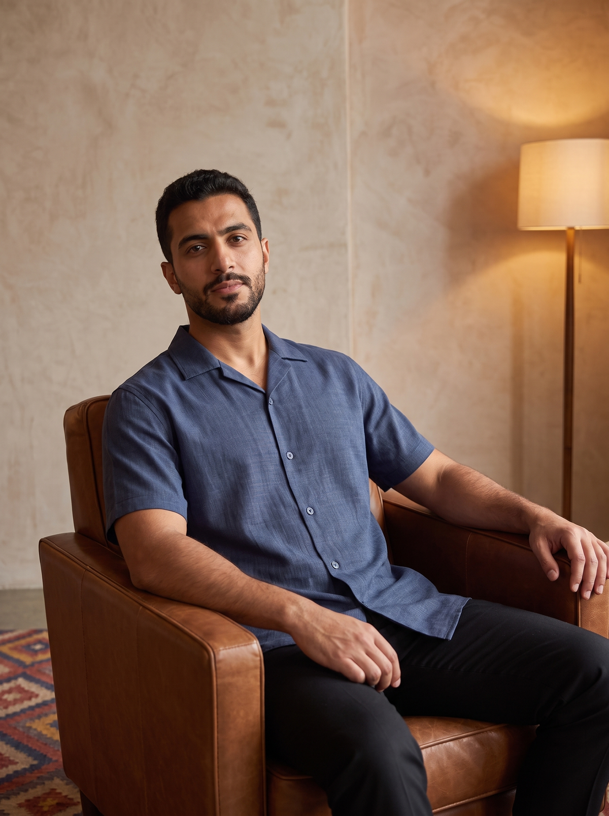 A man wearing a blue short-sleeve button-up shirt and black pants sits in a brown leather armchair in a warmly lit room with a textured beige wall and a floor lamp.