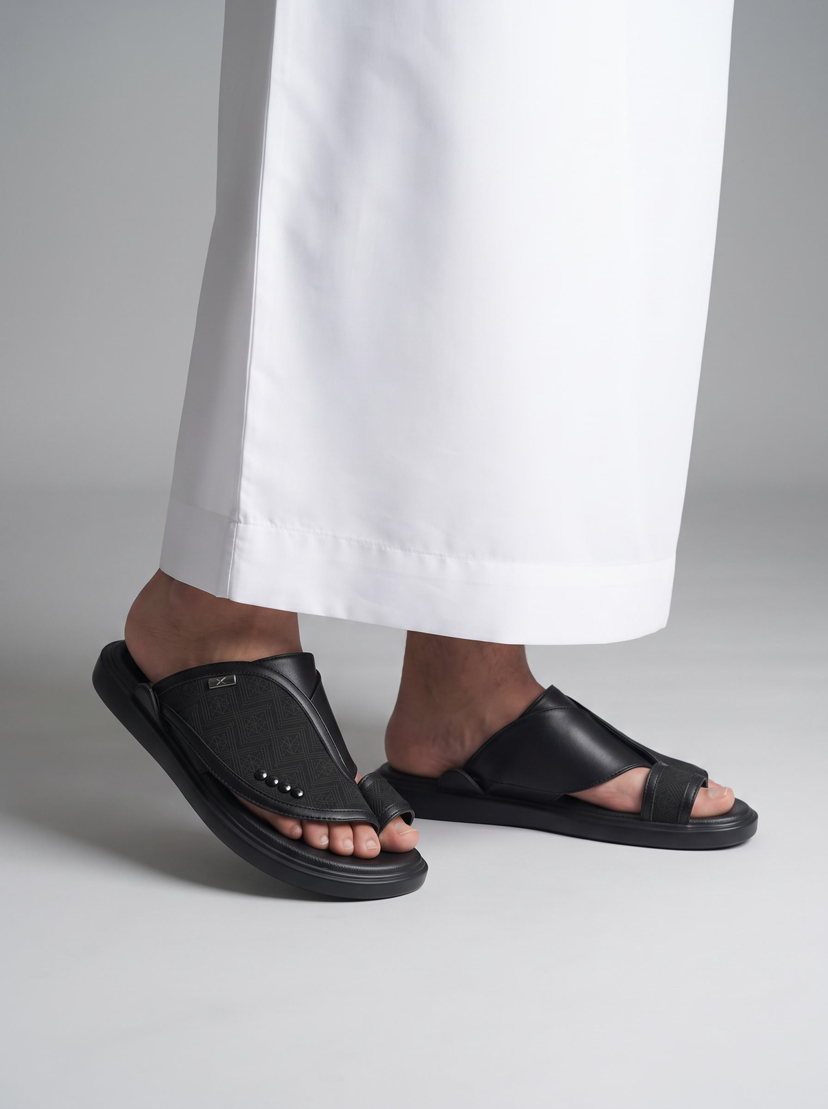 Black slide sandals worn with white wide-leg trousers, shown on a model's feet against a neutral gray studio background.