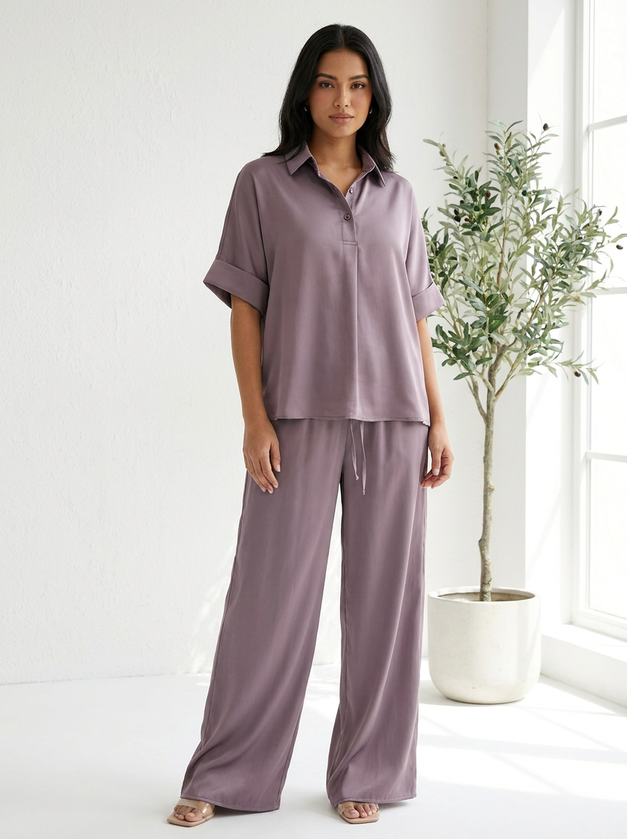 Woman wearing a mauve pajama set in a bright room with a plant.