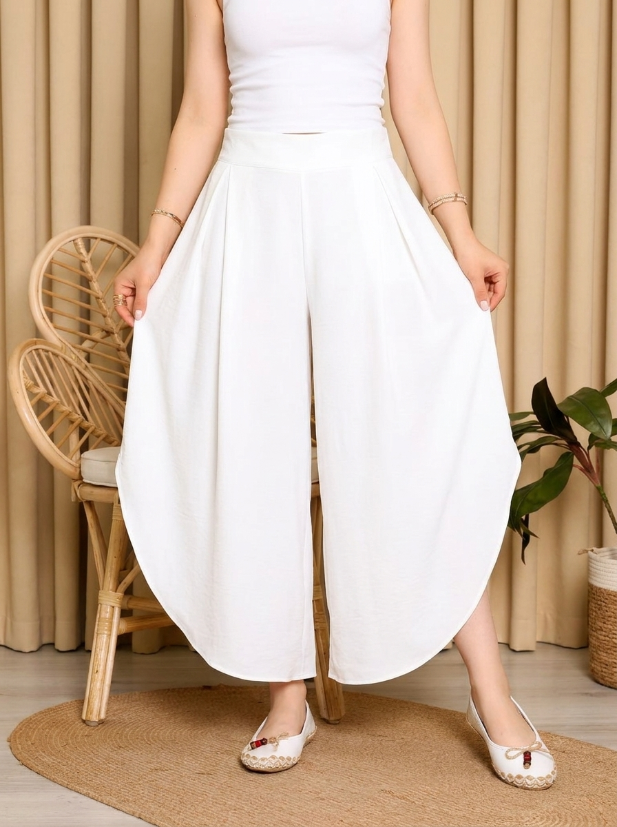 Person wearing a white dress with wide-leg pants in a room with beige curtains and a wicker chair.