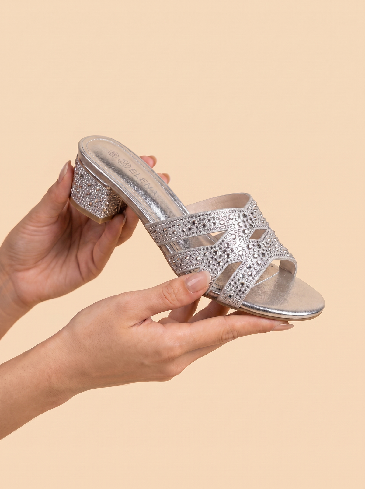 A pair of women's open-toe heeled mule sandals in metallic silver, held by hands against a solid beige background. The sandals feature intricate laser-cut geometric patterns across the straps and a textured, rhinestone-embellished block heel.