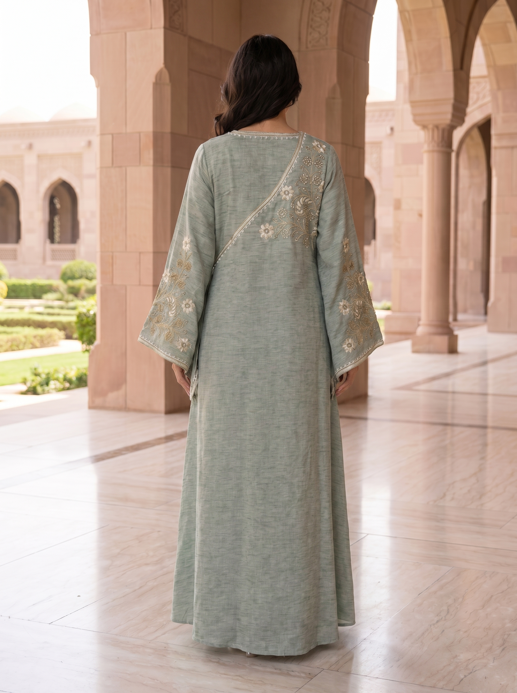 Woman in a long, light green dress with floral embroidery standing in an architectural setting.