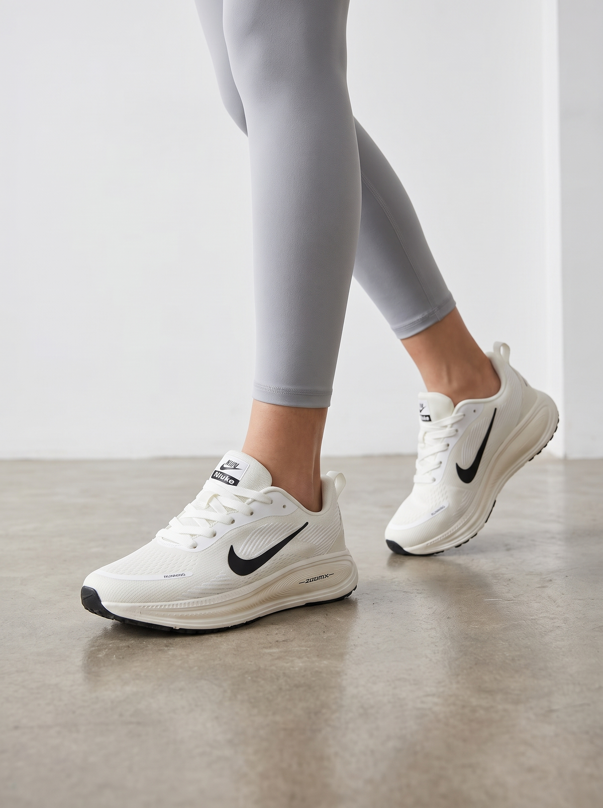 White Nike running shoes with black swooshes worn by a model in light gray leggings, shown from the knees down on a neutral indoor floor.