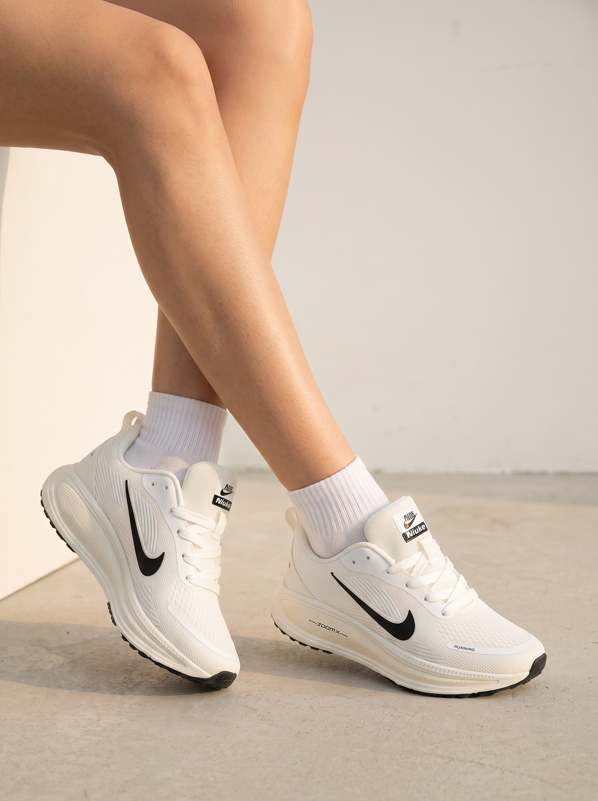 White Nike running shoes with black Swoosh logos, worn with white crew socks on a model's feet, shown against a minimalist concrete and wall background.