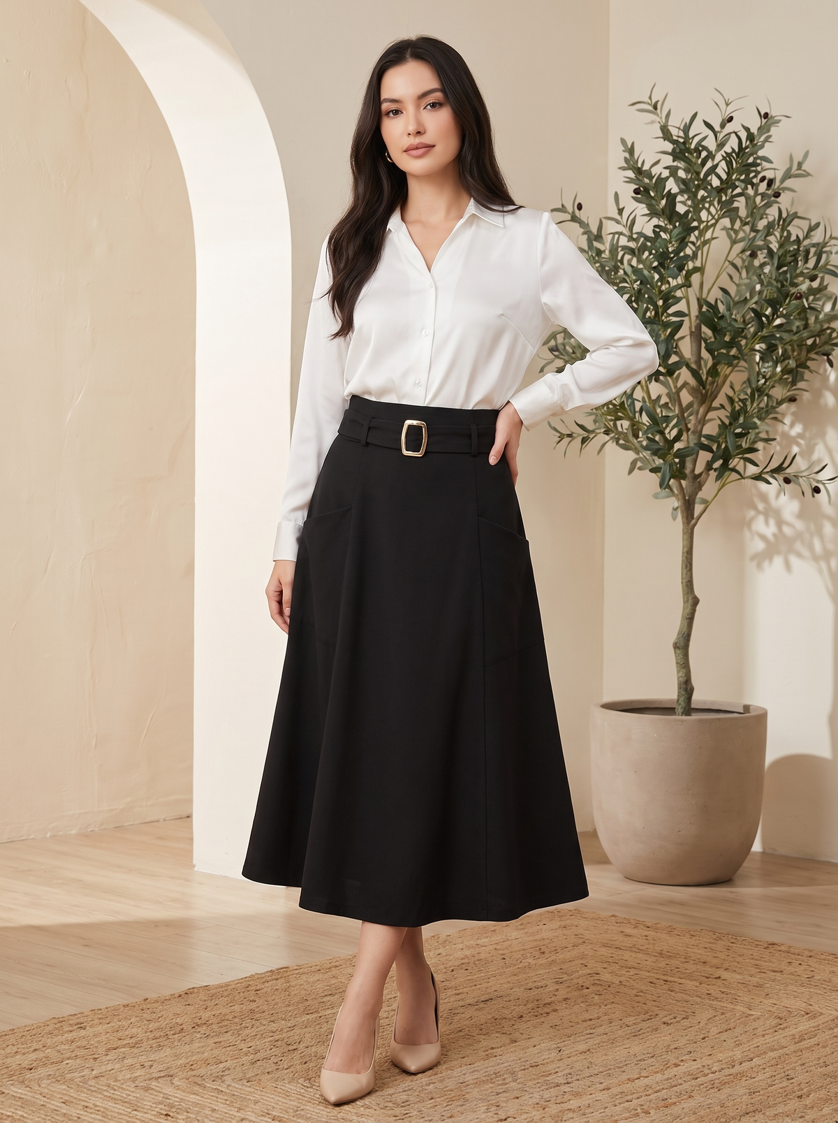 A woman modeling a black midi skirt with a matching belt and a white button-down shirt, standing in a warm, neutral-toned interior setting with a potted plant.