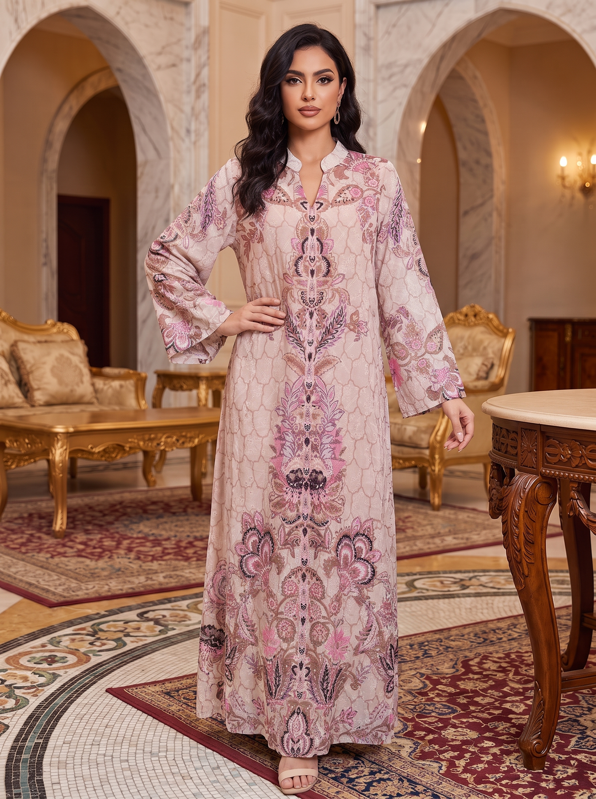 A woman models a long-sleeved, floor-length kaftan dress with an intricate pink, purple, and beige floral pattern, set in an elegant, ornate room with marble arches and traditional furniture.