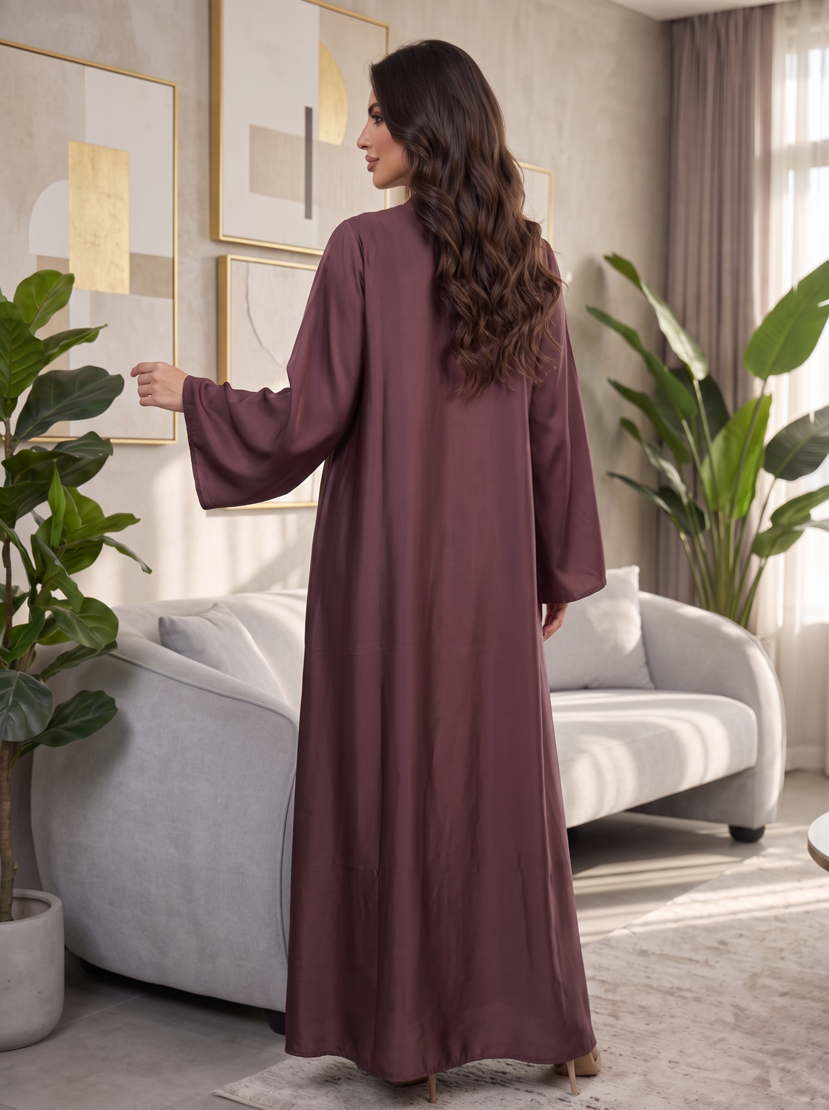 A woman wearing a long, loose-fitting, floor-length mauve-brown abaya with wide sleeves, standing in a modern living room with a gray sofa and potted plants.