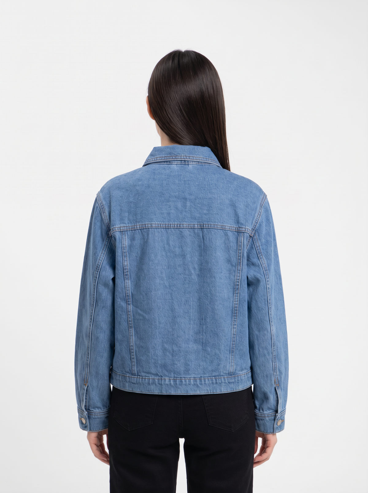 A woman viewed from the back wearing a light blue denim jacket and black pants against a plain white studio background.