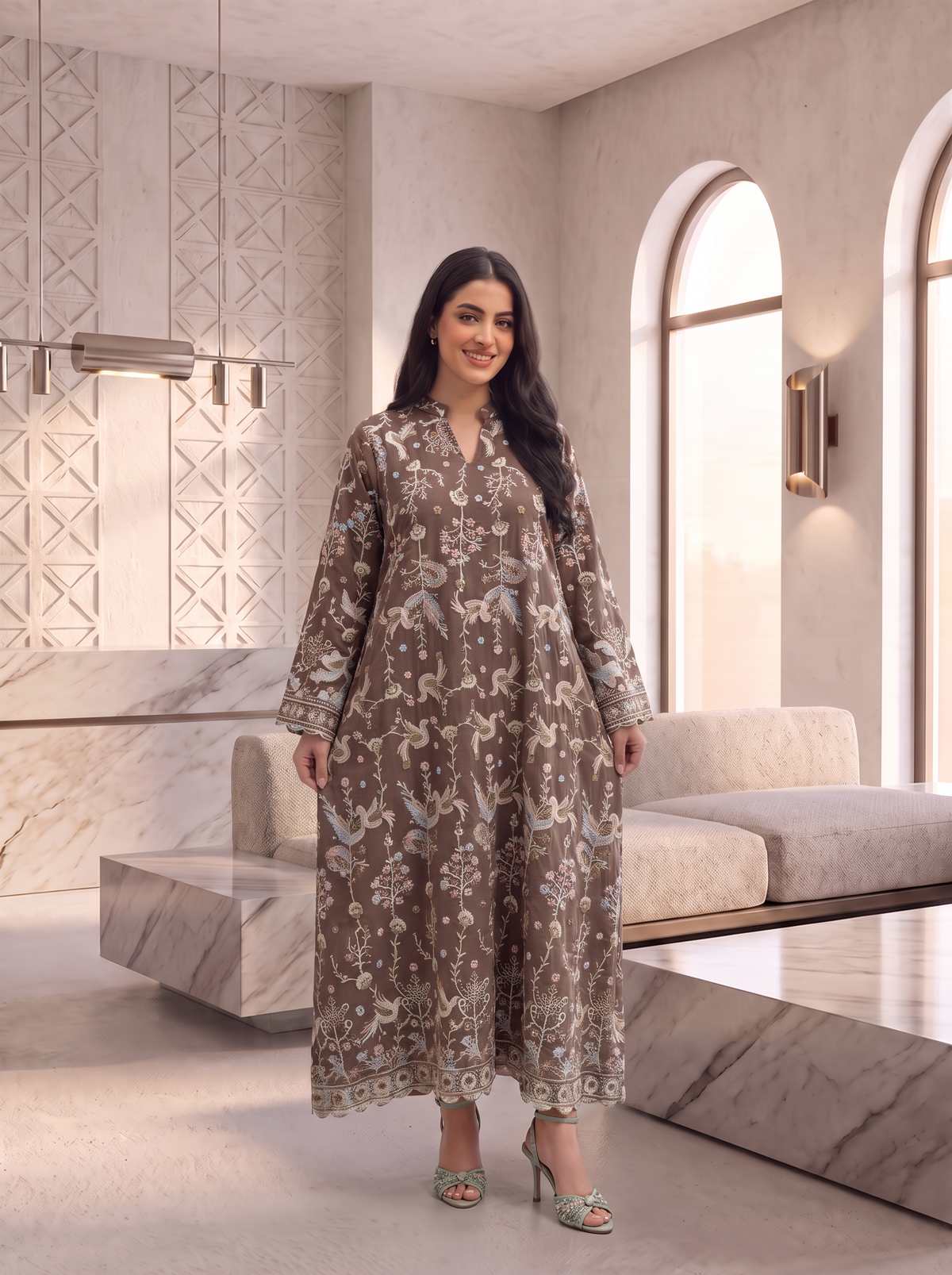 A woman modeling a long-sleeved, floor-length brown tunic dress with a beige and white floral pattern, standing in a modern, minimalist interior with marble furniture and arched windows.