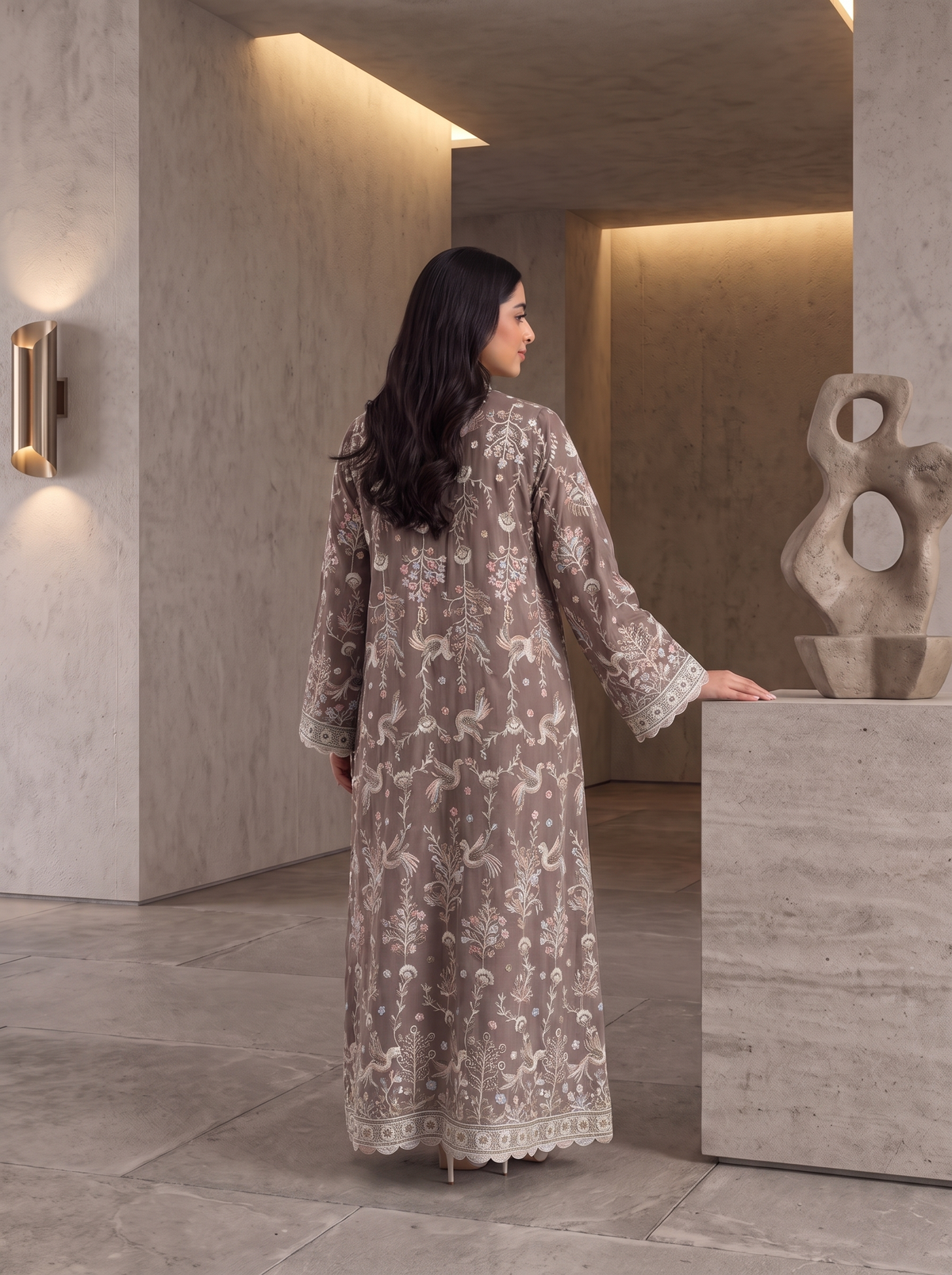 A woman stands with her back to the camera wearing a long, beige and brown embroidered traditional dress in a modern, minimalist concrete-walled interior.