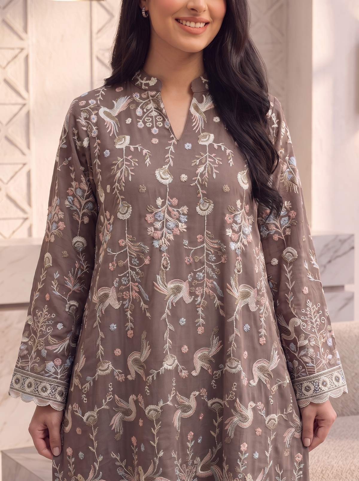 A woman wearing a brown long-sleeve kurta with intricate floral and bird embroidery, shown from the chest down in a softly lit indoor setting.