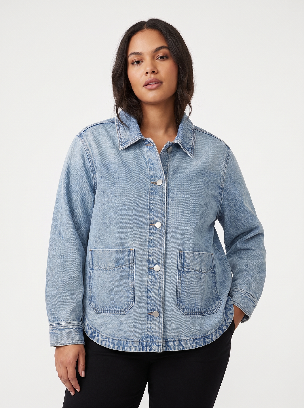 A woman modeling a light blue denim button-up shirt jacket with front pockets, paired with black pants, against a plain light gray studio background.