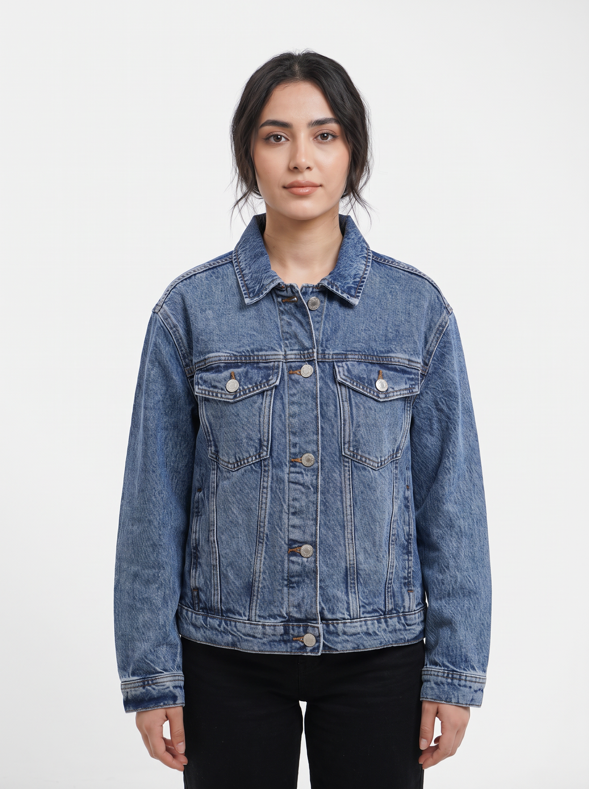A woman modeling a classic blue denim jacket with front button closure, two buttoned chest pockets, and a pointed collar, paired with black pants against a plain light gray background.