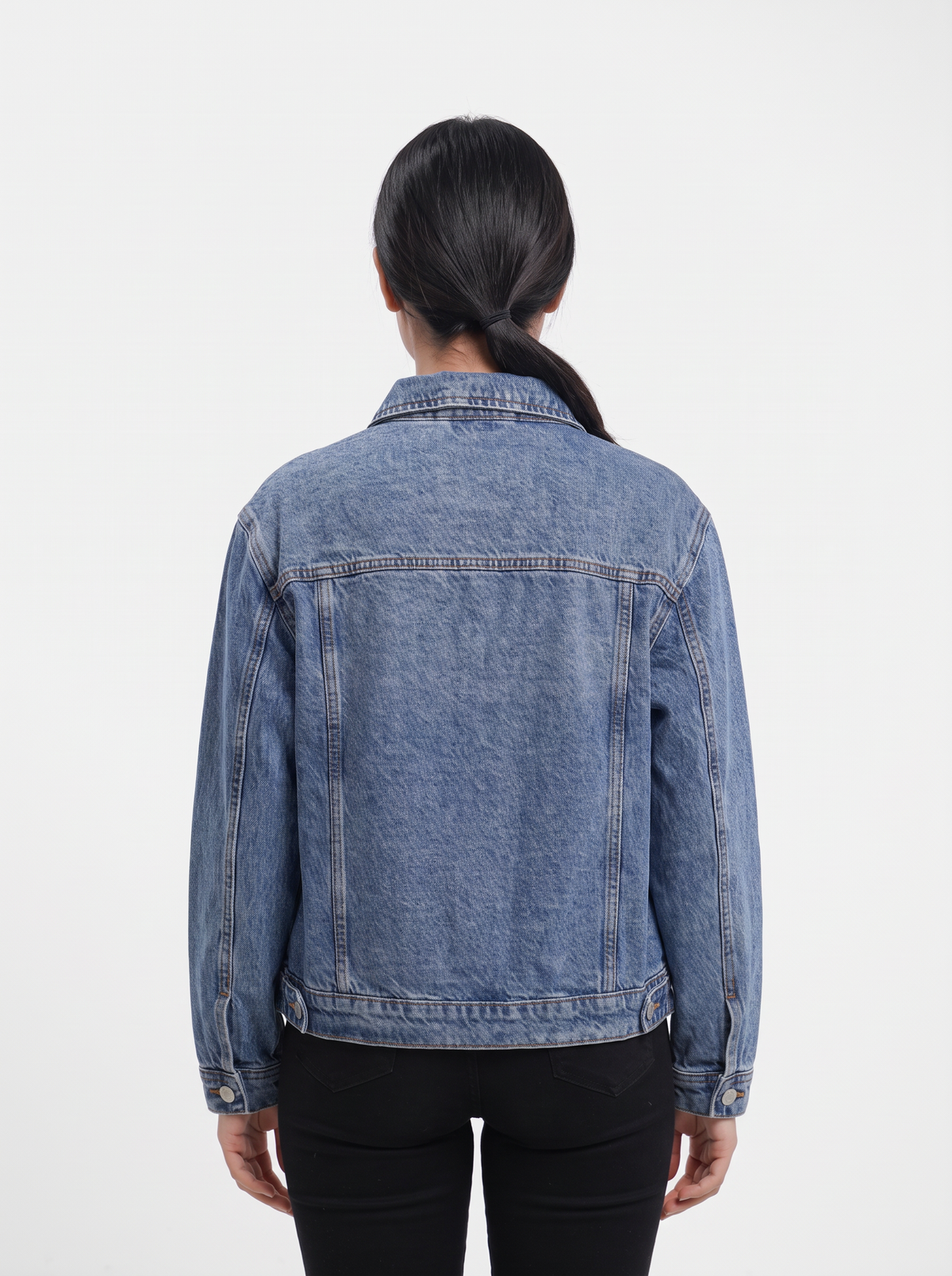 A person viewed from the back wearing a light-to-medium wash denim jacket over a black top, against a plain light gray studio background.