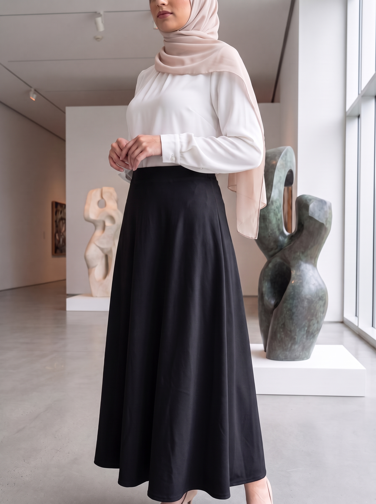 A woman wearing a white long-sleeve blouse and a high-waisted black A-line midi skirt, paired with a light beige hijab, standing in a modern art gallery with abstract sculptures.