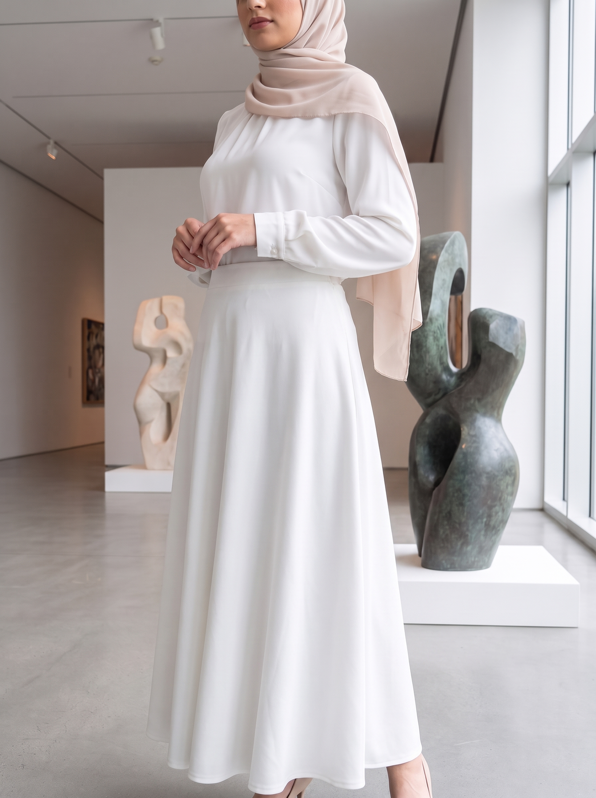 A woman wearing a long-sleeve white maxi dress and a light beige hijab stands in a modern art gallery, with abstract sculptures visible in the background.