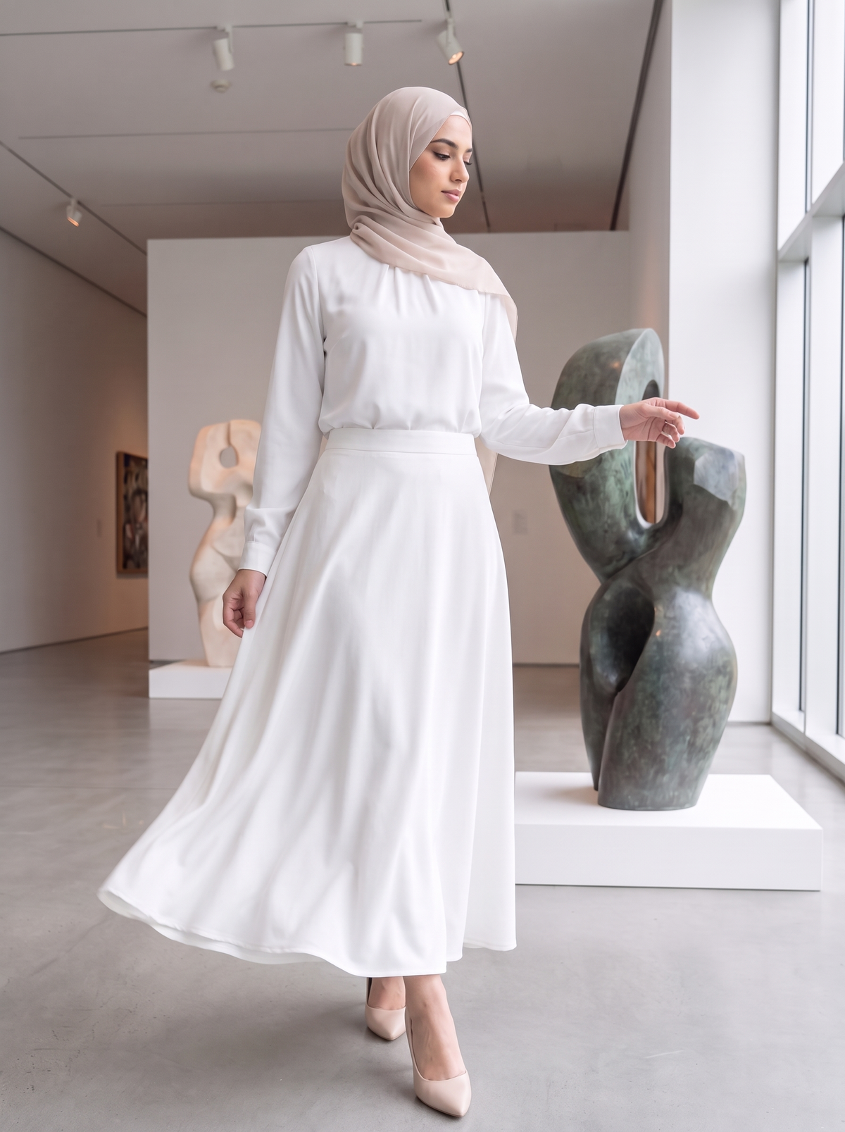 A woman wearing a white long-sleeve maxi dress and a light beige hijab stands in a modern art gallery, gesturing toward a bronze abstract sculpture.