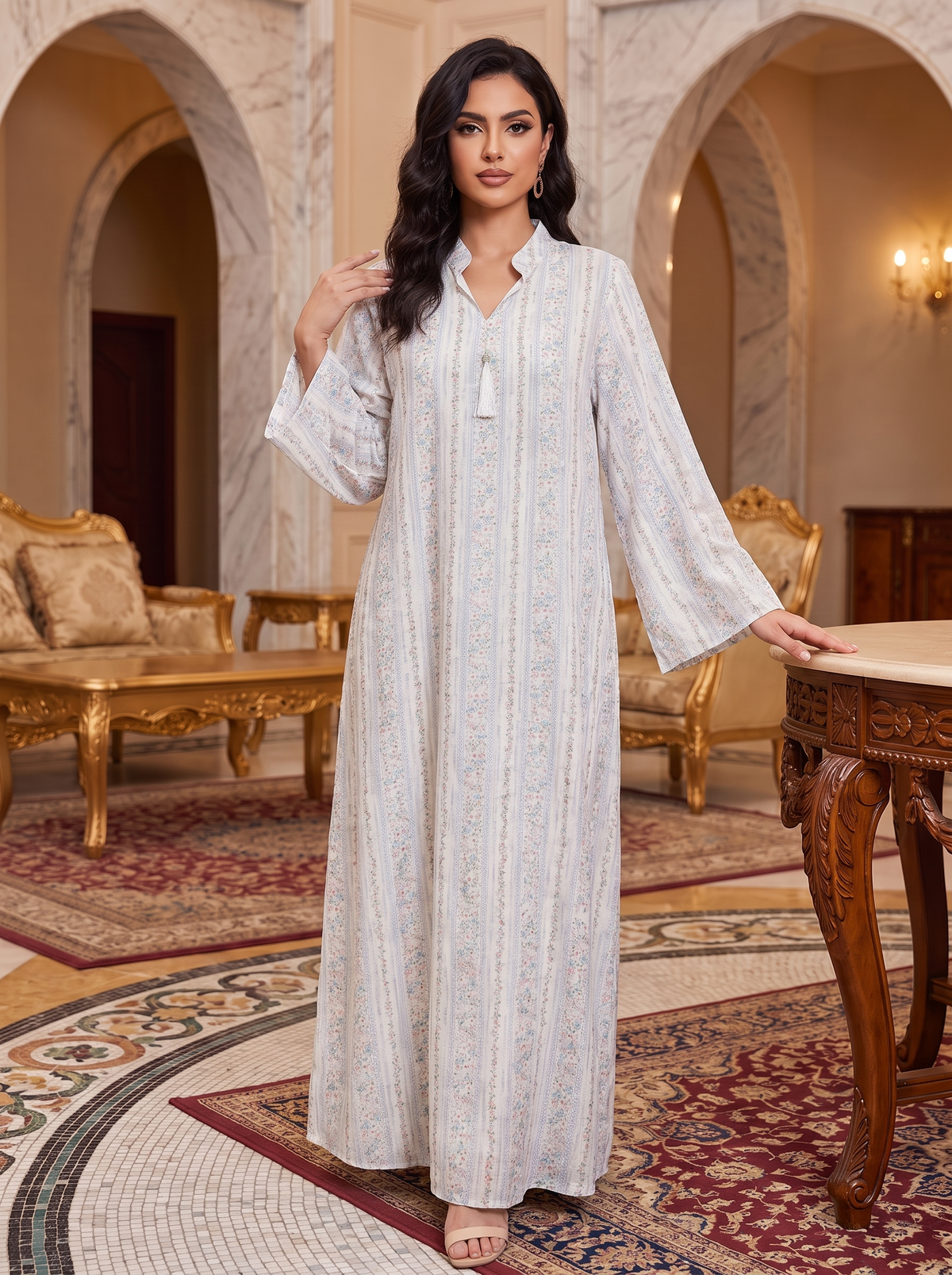 A woman models a long, flowing kaftan dress in a luxurious, ornate room with marble arches and gold furniture. The dress is off-white with vertical stripes and delicate floral embroidery.