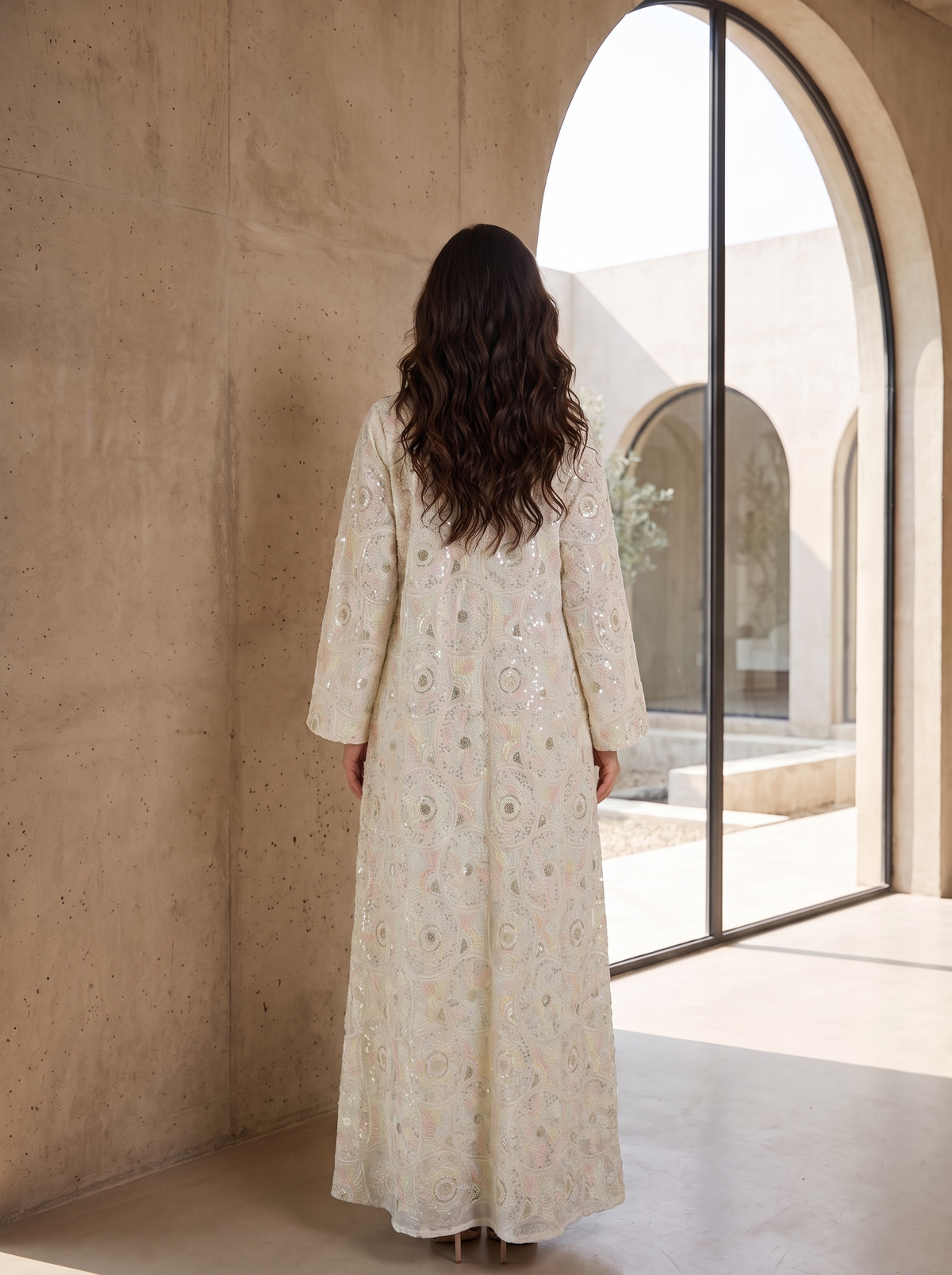 A woman viewed from behind wearing a long cream-colored embroidered caftan with intricate multicolor floral patterns, standing in a sunlit architectural space with concrete walls and a large arched window.