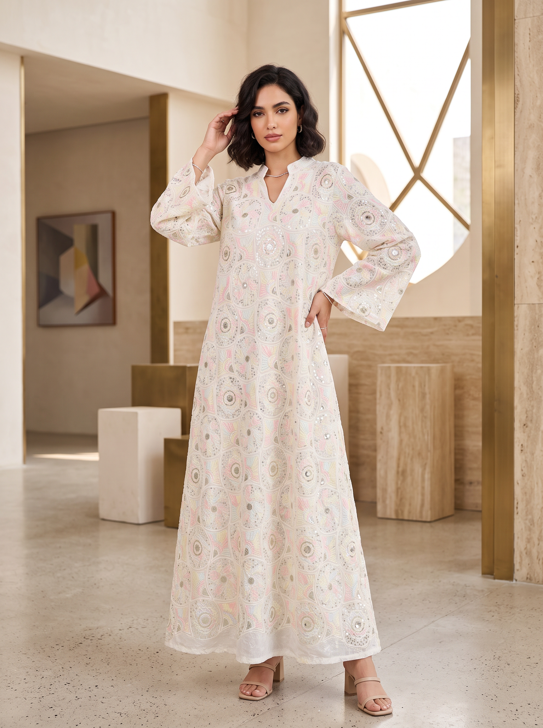 A woman modeling a long white maxi dress with delicate pink and green floral embroidery, standing in a modern, sunlit interior space.