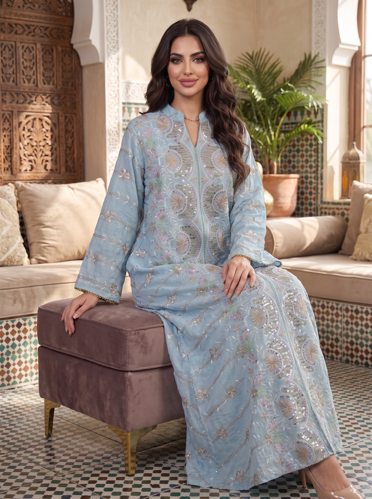 A woman wearing a light blue embroidered kaftan robe sits on a brown velvet ottoman in a Moroccan-style room with patterned tile floors and cushions.