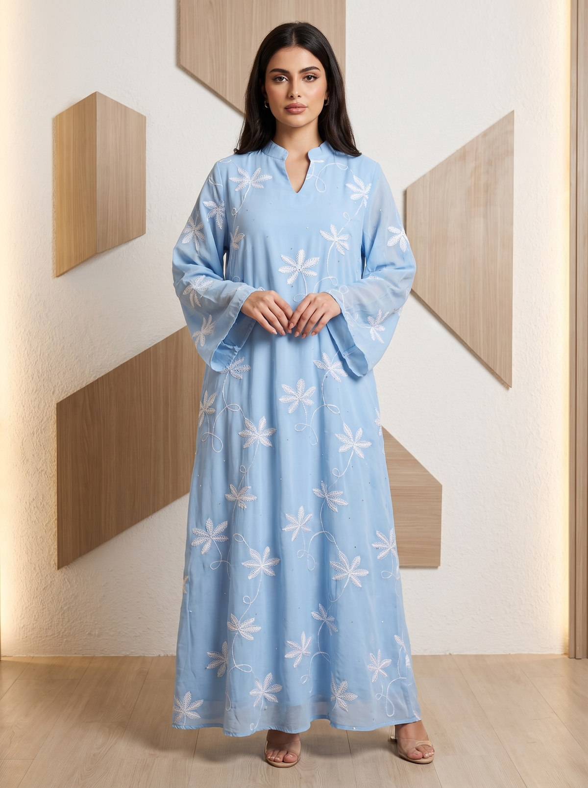 A woman stands indoors wearing a light blue long kaftan-style dress with white floral embroidery and wide sleeves. The room has a cream textured wall with geometric wood panel accents and light wood flooring.