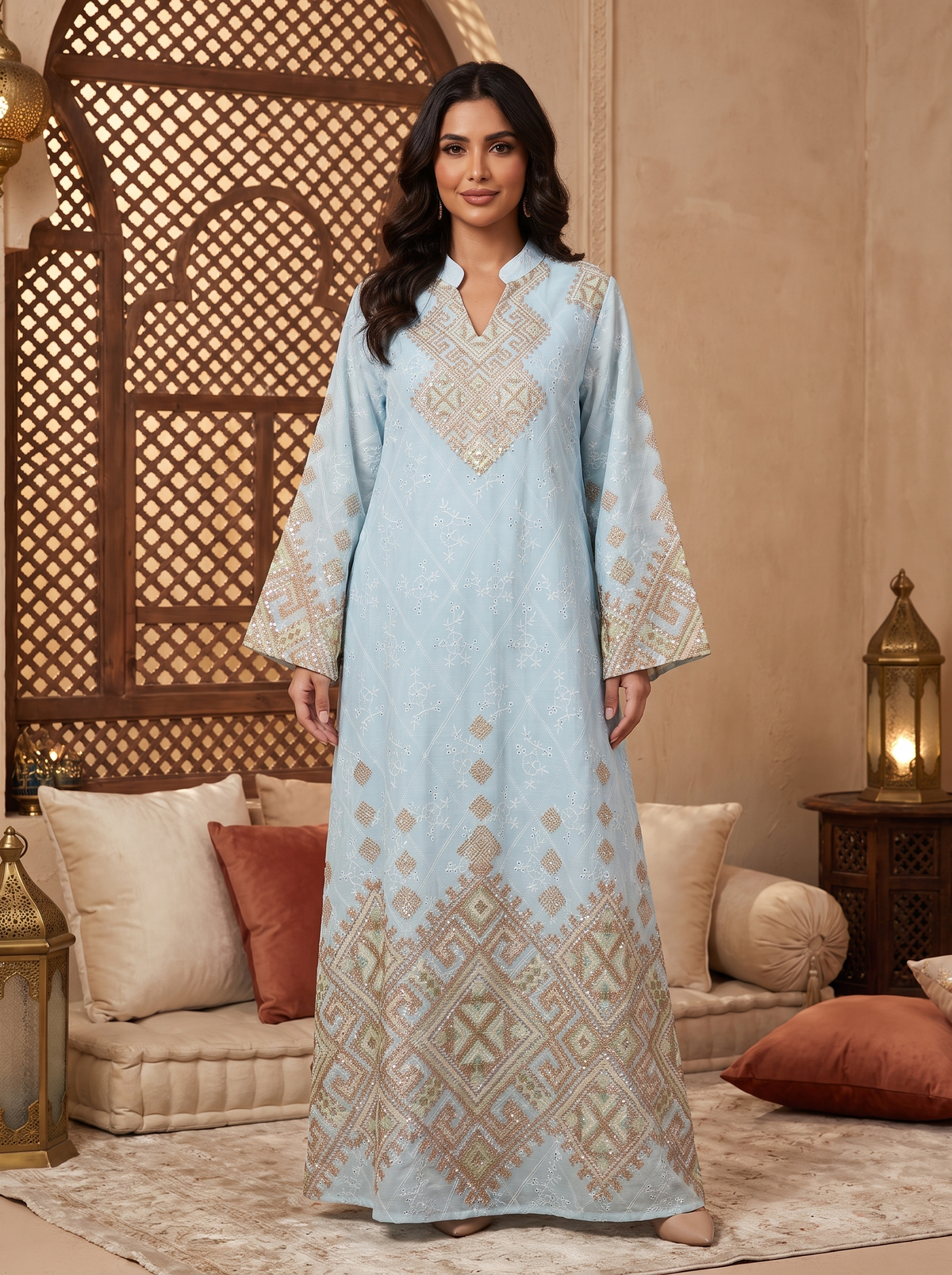 A woman stands in a decorated indoor setting wearing a long light blue embroidered dress with beige and gold geometric patterns. The room features a beige wall, a wooden lattice screen, cushions, and lanterns.