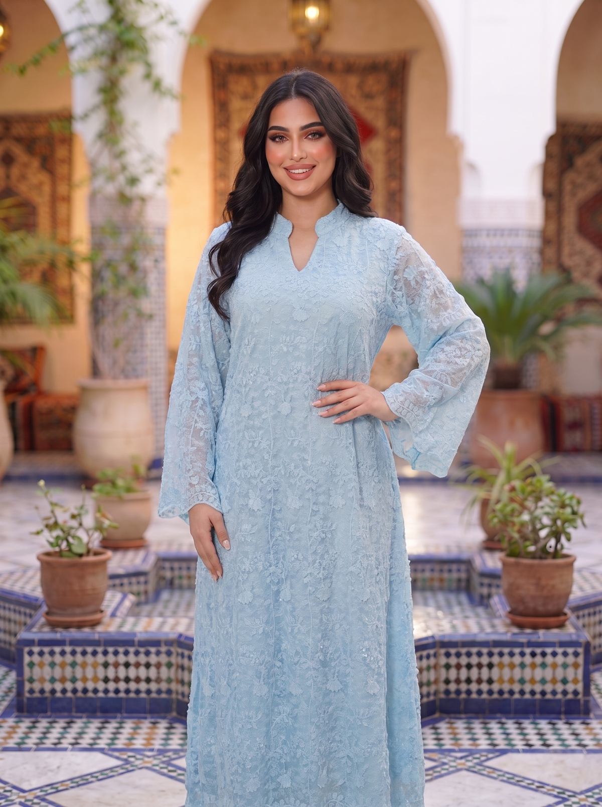 A woman modeling a light blue lace long-sleeve kaftan dress in an ornate Moroccan-style courtyard with tiled floors and potted plants.