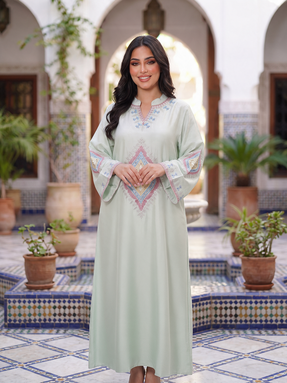 A woman stands in a tiled courtyard wearing a long, light sage green caftan with colorful embroidered details on the chest and sleeves. The background features white arches, potted plants, and blue-and-white mosaic tile patterns.