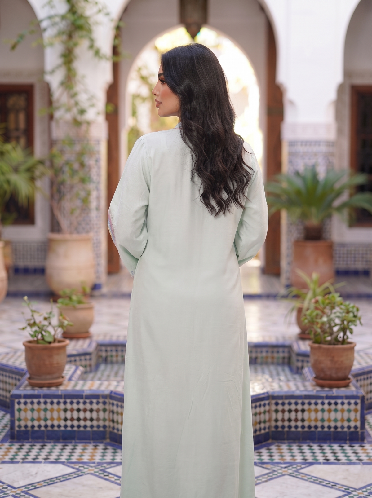 A woman with long dark wavy hair is shown from behind wearing a light sage green kaftan dress in an elegant Moroccan courtyard with blue and white patterned tile floors and arches.