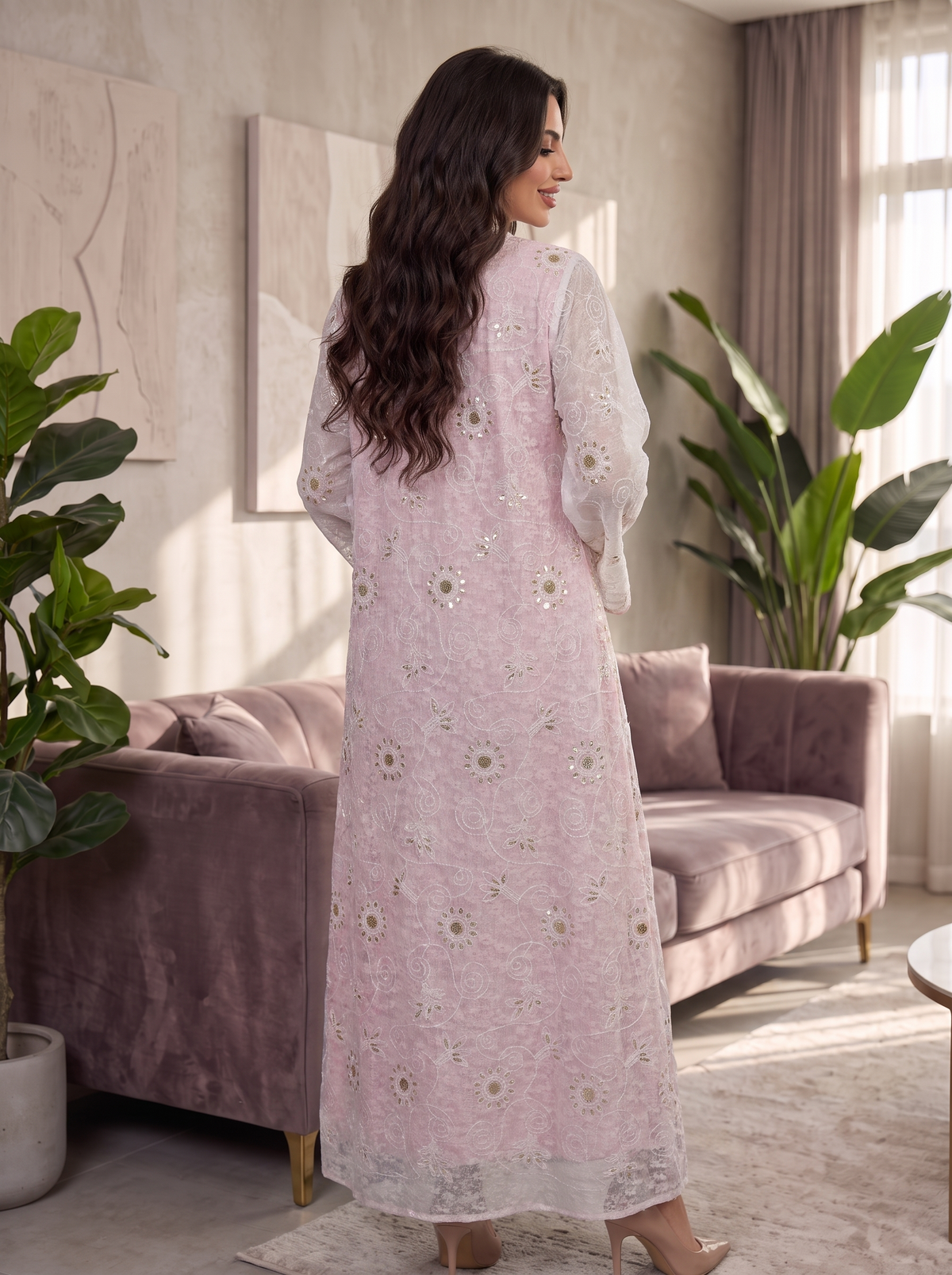 A woman stands in a living room wearing a long light pink embroidered dress or kaftan, photographed from the back. The garment has sheer sleeves and scattered floral embroidery in pale pink, white, and beige.