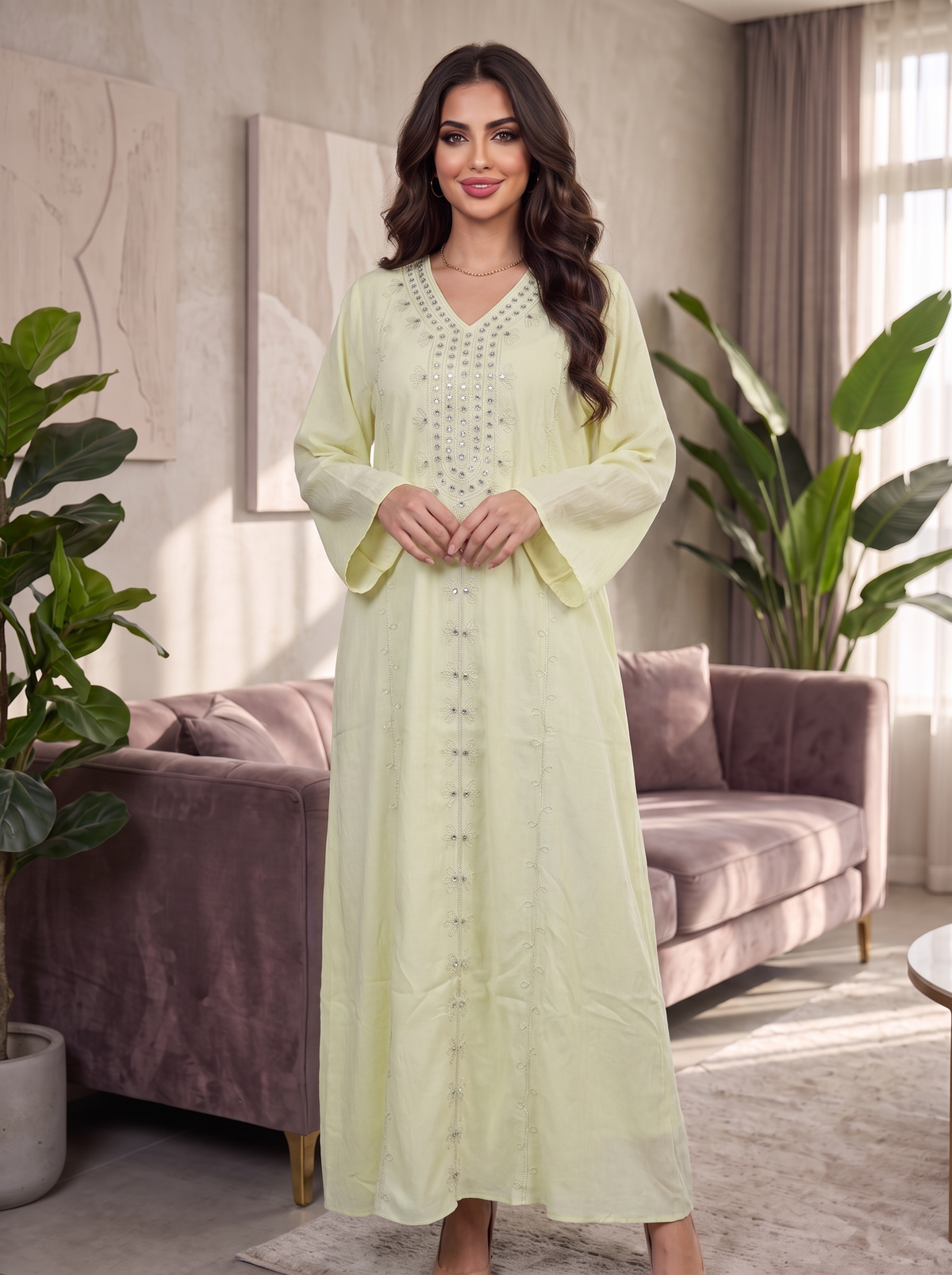 A woman stands in a modern living room wearing a long, light yellow-green kaftan dress with silver embellishments down the front and on the neckline. The dress features wide bell sleeves and a V-neckline.