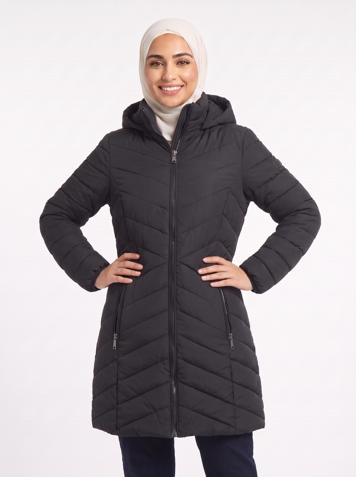 A woman wearing a black quilted hooded puffer coat with a front zipper and zip pockets, paired with a light beige hijab, posing against a solid white studio background.