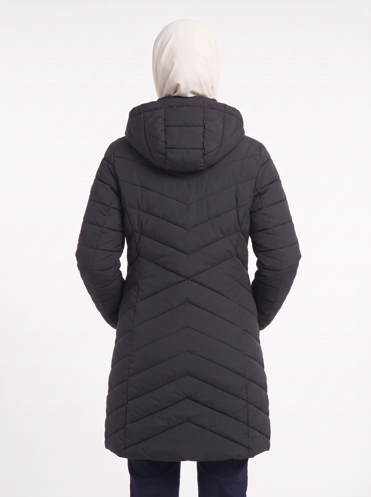 A person shown from the back wearing a long black quilted puffer coat with a hood and a white hijab, against a plain white studio background.