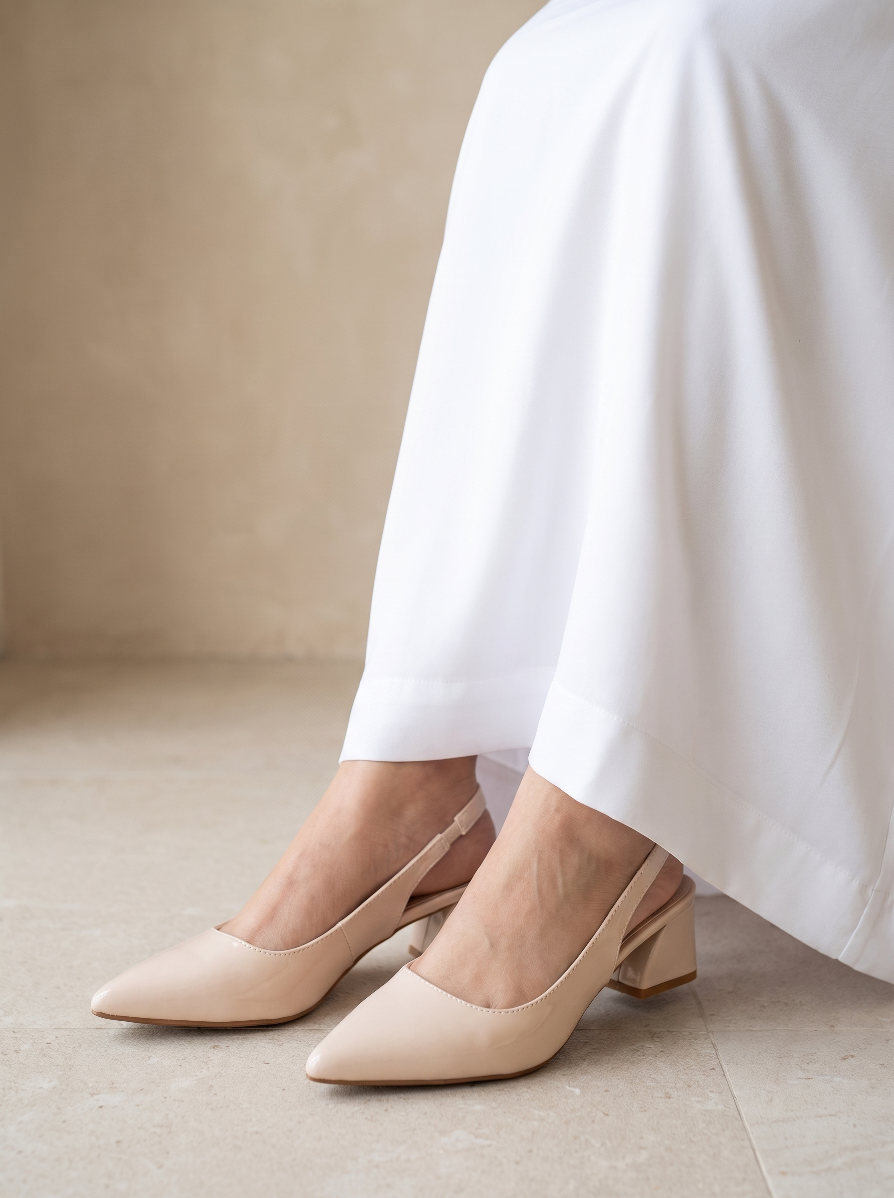 A pair of beige pointed-toe slingback pumps with a low block heel, shown on a model wearing white draped pants against a neutral beige background.