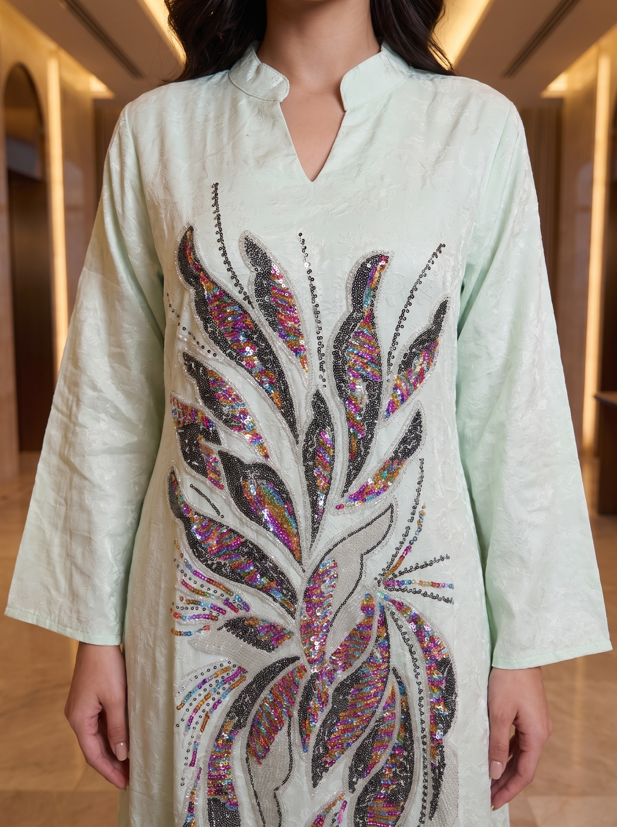 A woman wearing a light sage green kurta with a large, colorful sequined leaf design on the front, set against a blurred indoor hallway background.