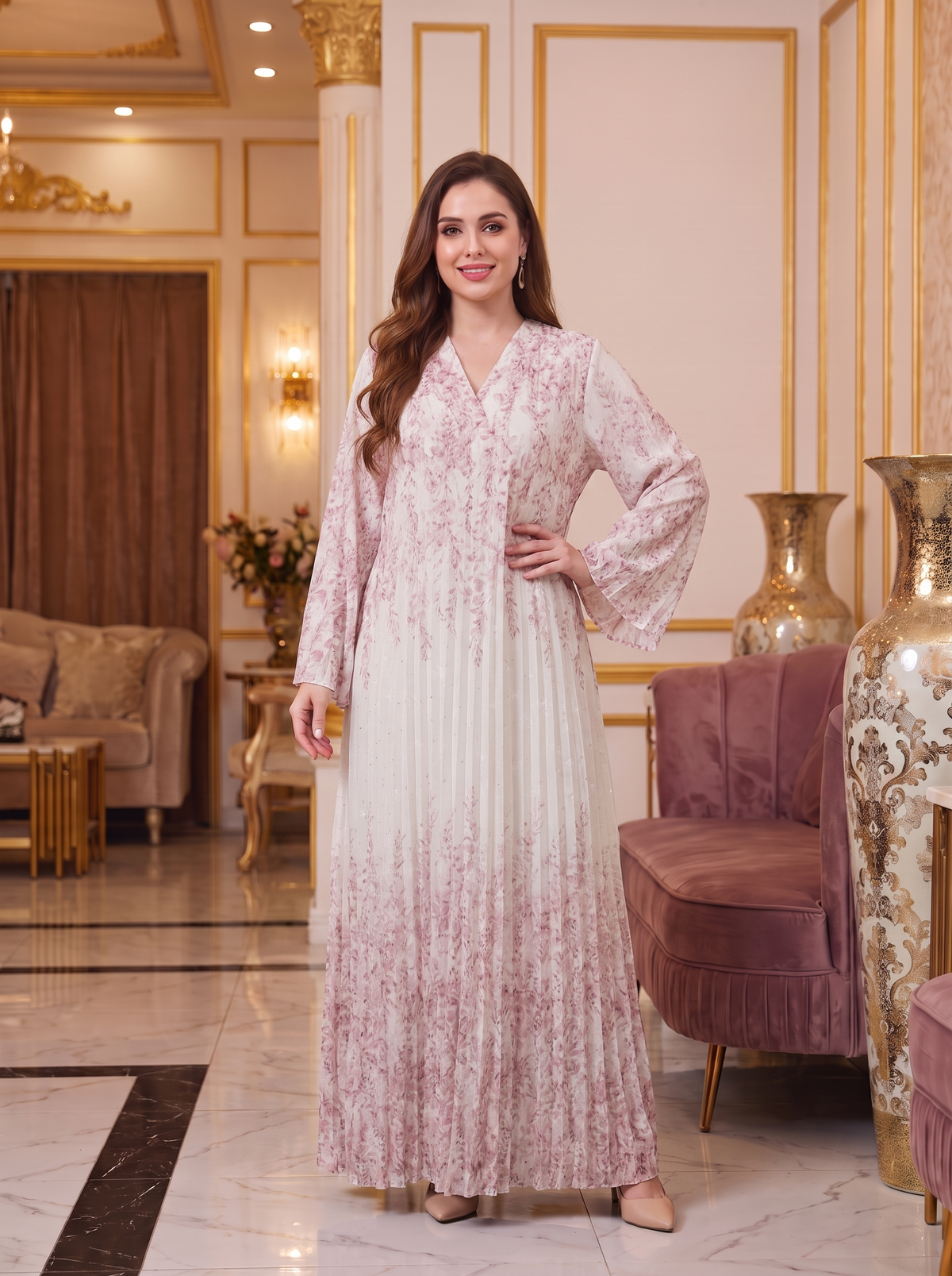 A woman stands in a luxurious interior wearing a long, flowing maxi dress with a light pink and white floral pattern. The dress features a V-neckline, long flared sleeves, and a pleated skirt.