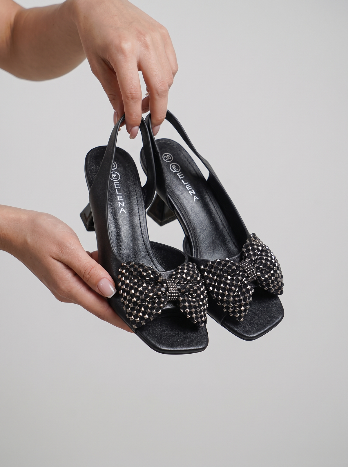 Black slingback block-heel sandals with large studded bow details on the toes, held by hands against a light gray background.