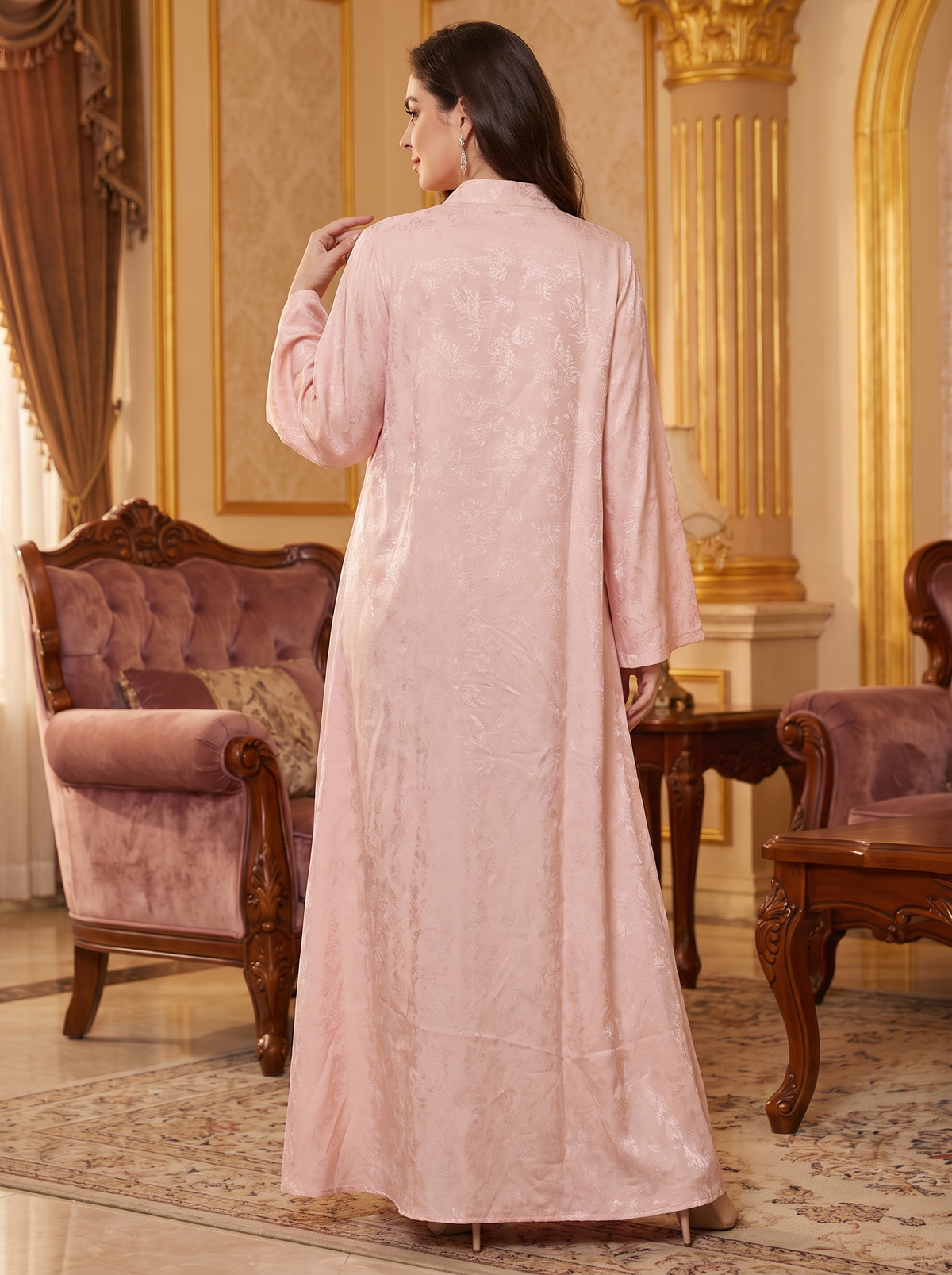 A woman stands with her back to the camera wearing a long, light pink embroidered abaya in an ornate, luxurious room with gold columns and purple velvet armchairs.
