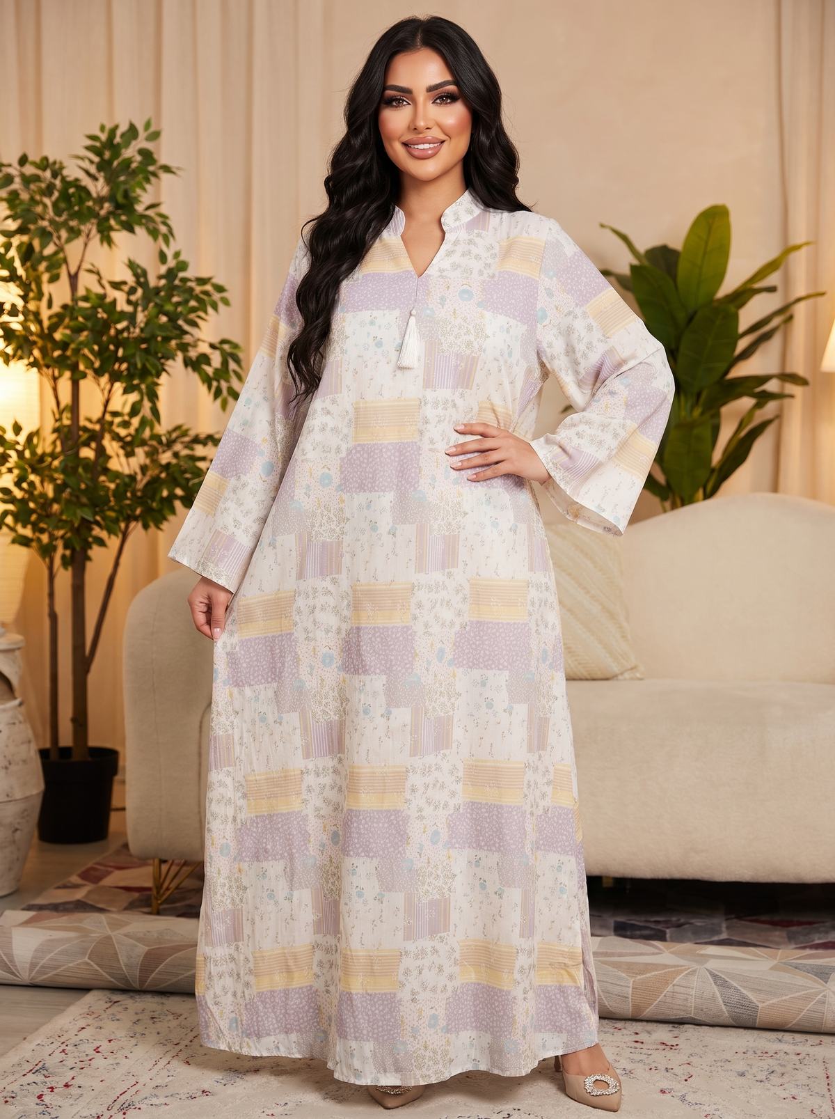 A woman modeling a long, loose patchwork kaftan dress with a V-neckline and wide bell sleeves, featuring a mix of lavender, beige, and white patterns, in a cozy living room setting.