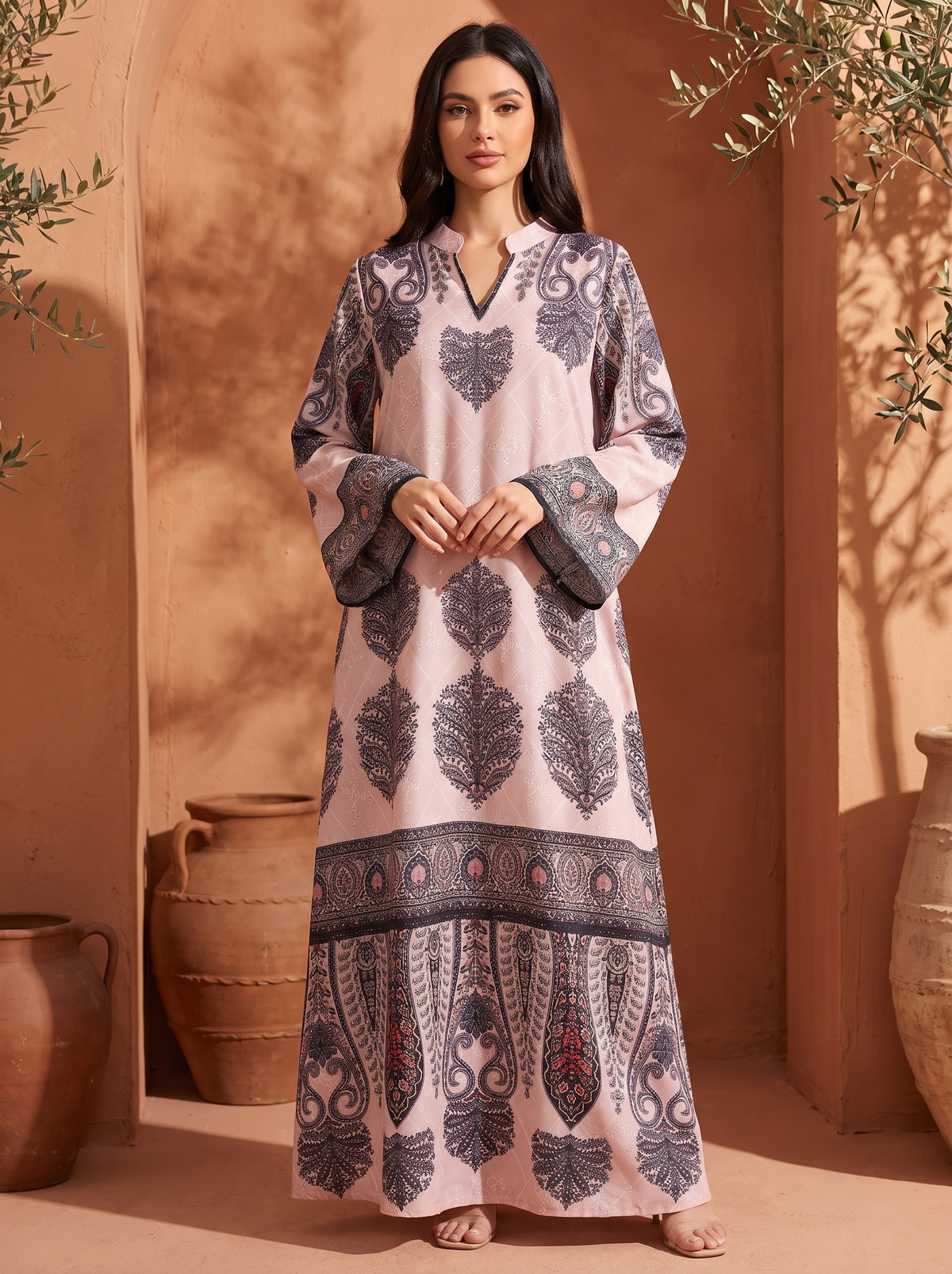 A woman stands outdoors wearing a long light pink kaftan dress with dark gray and black floral motifs and a decorative border. The setting features warm terracotta walls, large clay pots, and leafy branches.