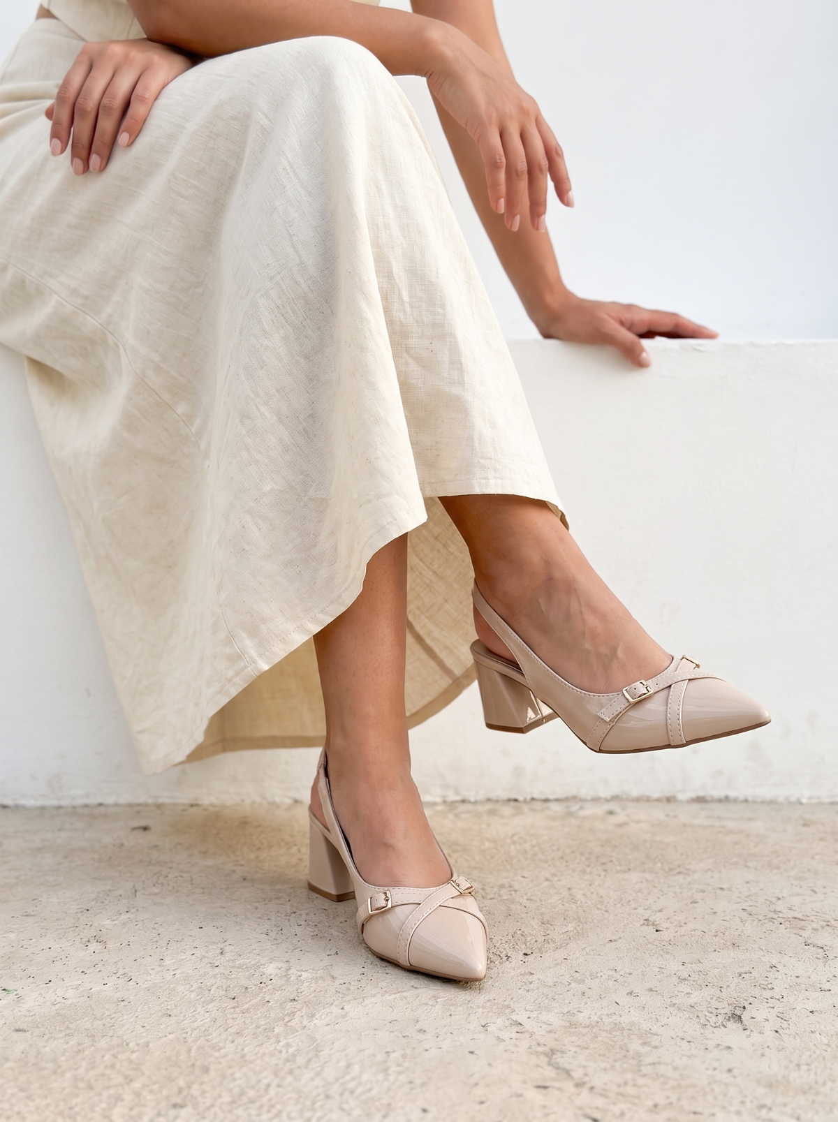 A person seated on a white ledge wearing light beige slingback block-heel pumps with small buckle details, paired with a flowing cream dress. The photo is styled in a bright, minimalist setting with a concrete floor.