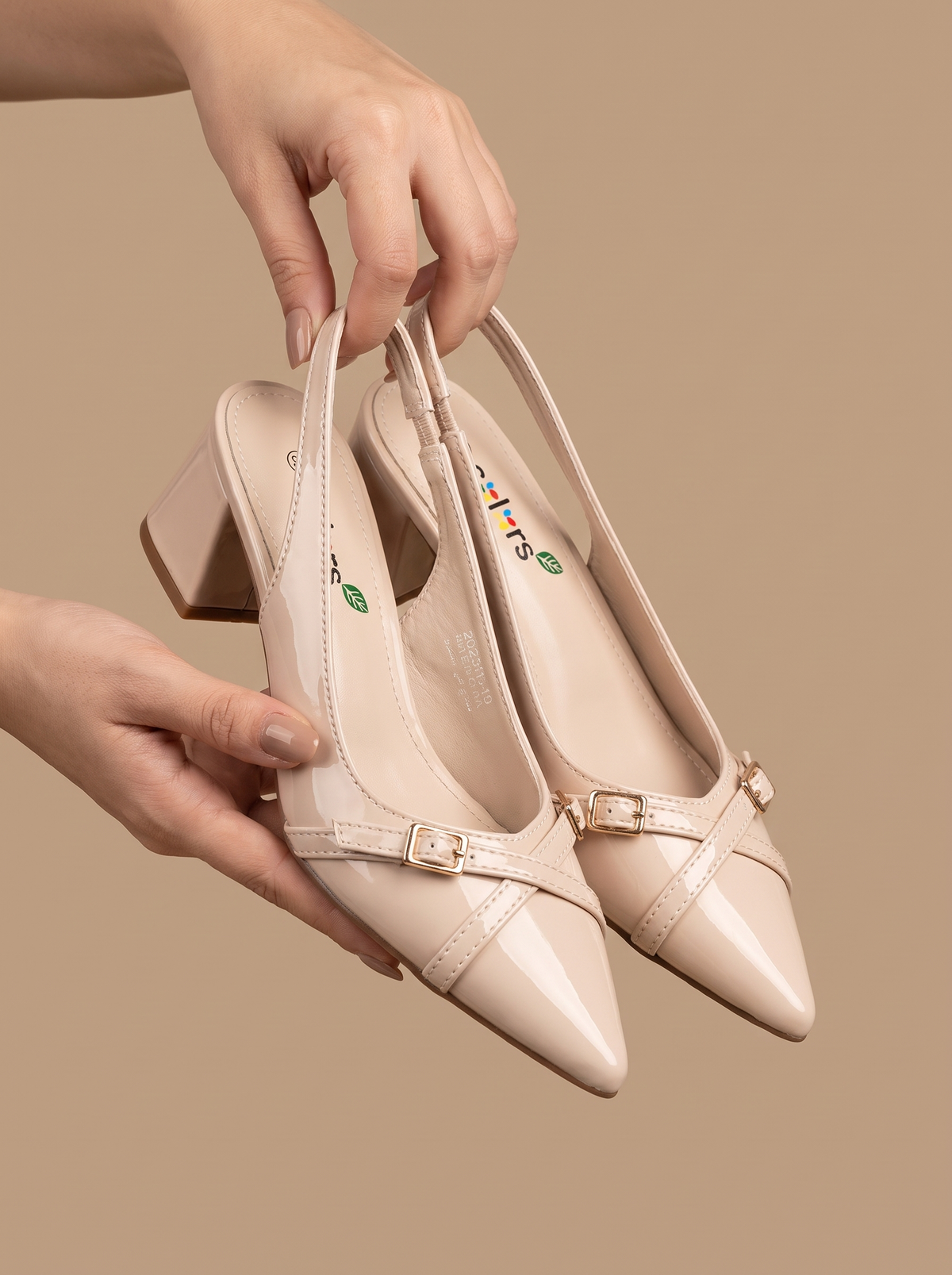 A pair of beige pointed-toe slingback pumps with gold buckle details, held by hands against a solid beige background.