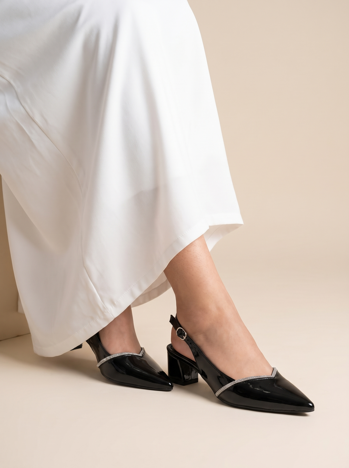 A person wearing black pointed-toe slingback heels with a silver metallic trim accent, paired with a flowing white skirt, shown against a solid beige studio background.