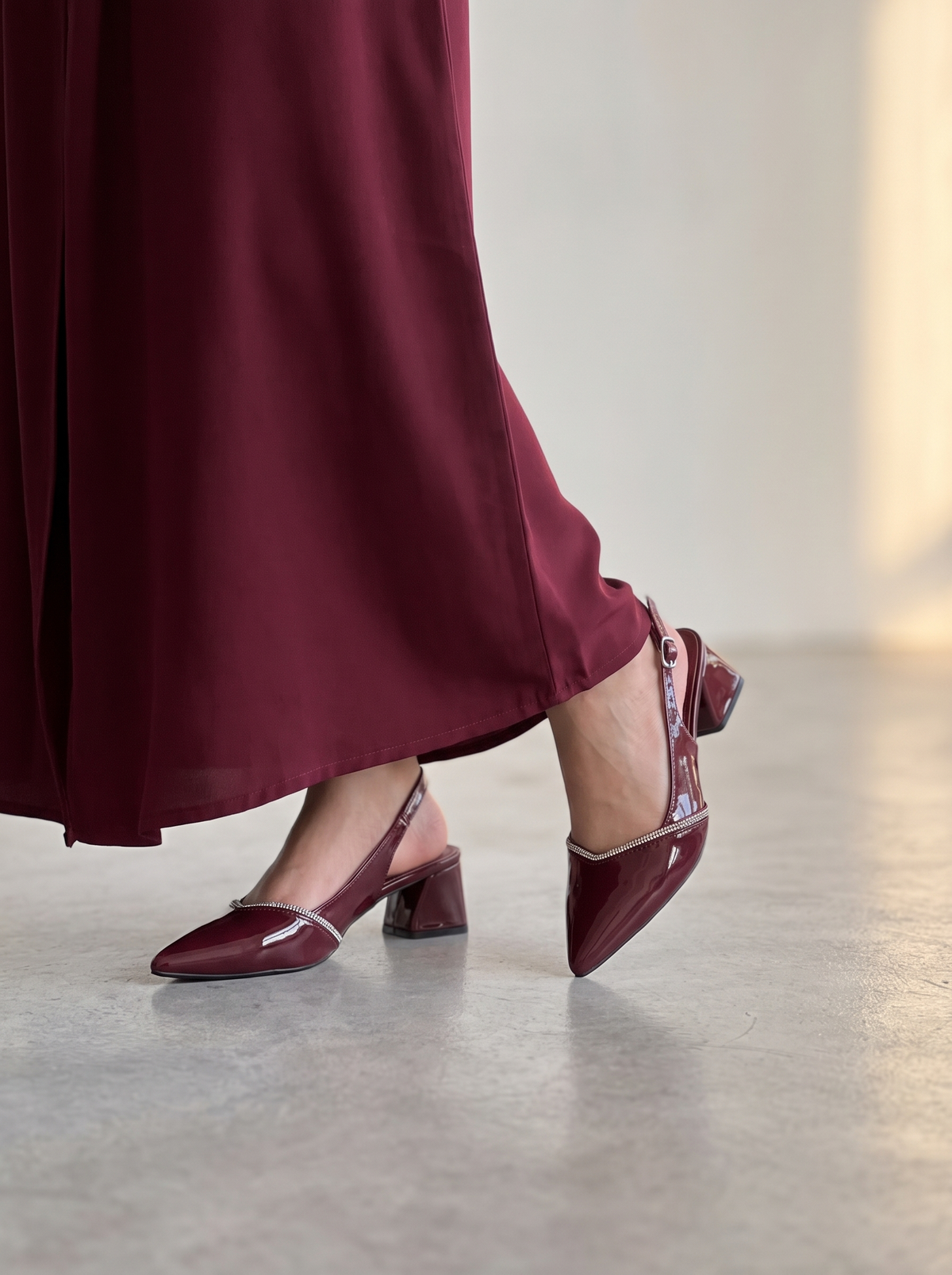 A person wearing burgundy pointed-toe slingback heels with a block heel and a matching long burgundy skirt, photographed in a softly lit indoor setting with a concrete floor.