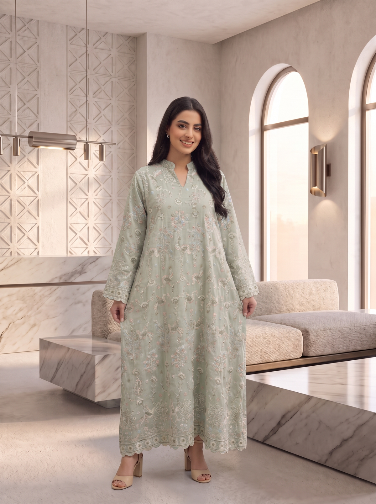 A woman modeling a long, light sage green embroidered kurta in a modern, elegant living room setting with marble furniture and arched windows.
