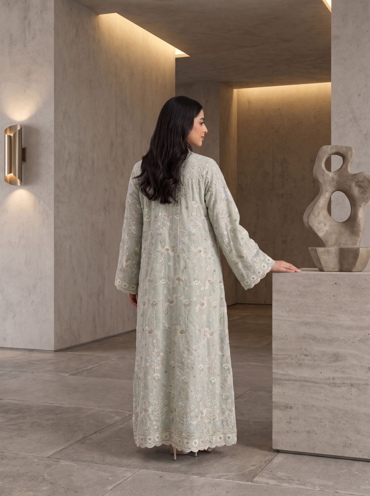 A woman wearing a long, light sage green embroidered kaftan dress stands in a modern minimalist interior with concrete walls and warm lighting.