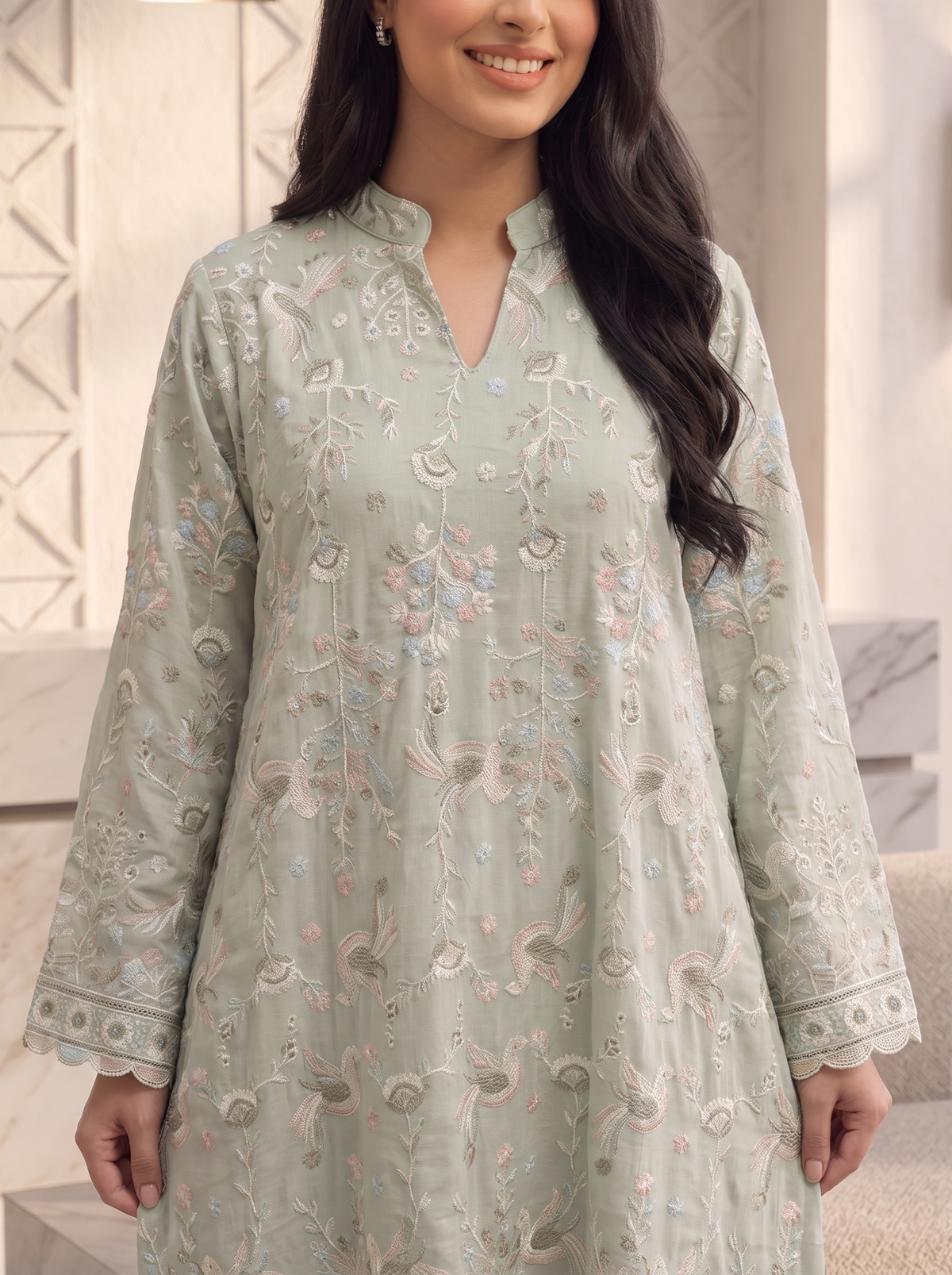 A woman wearing a light sage green embroidered kurta with floral and paisley patterns in beige, pink, and blue, shown from the chest down in a softly lit indoor setting.