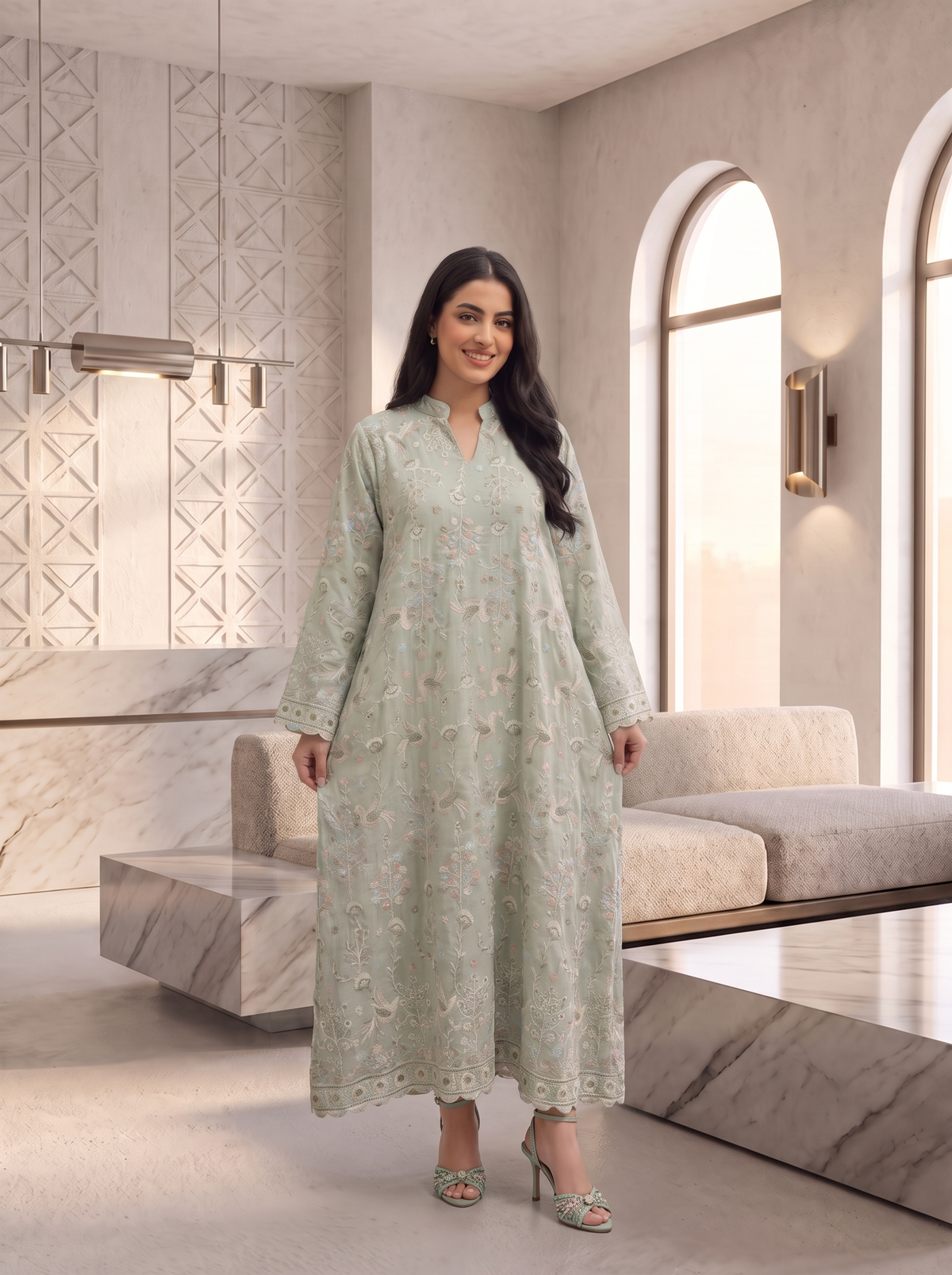 A woman modeling a long, light sage green embroidered kurta with floral patterns, standing in a modern, minimalist living room with marble accents and arched windows.