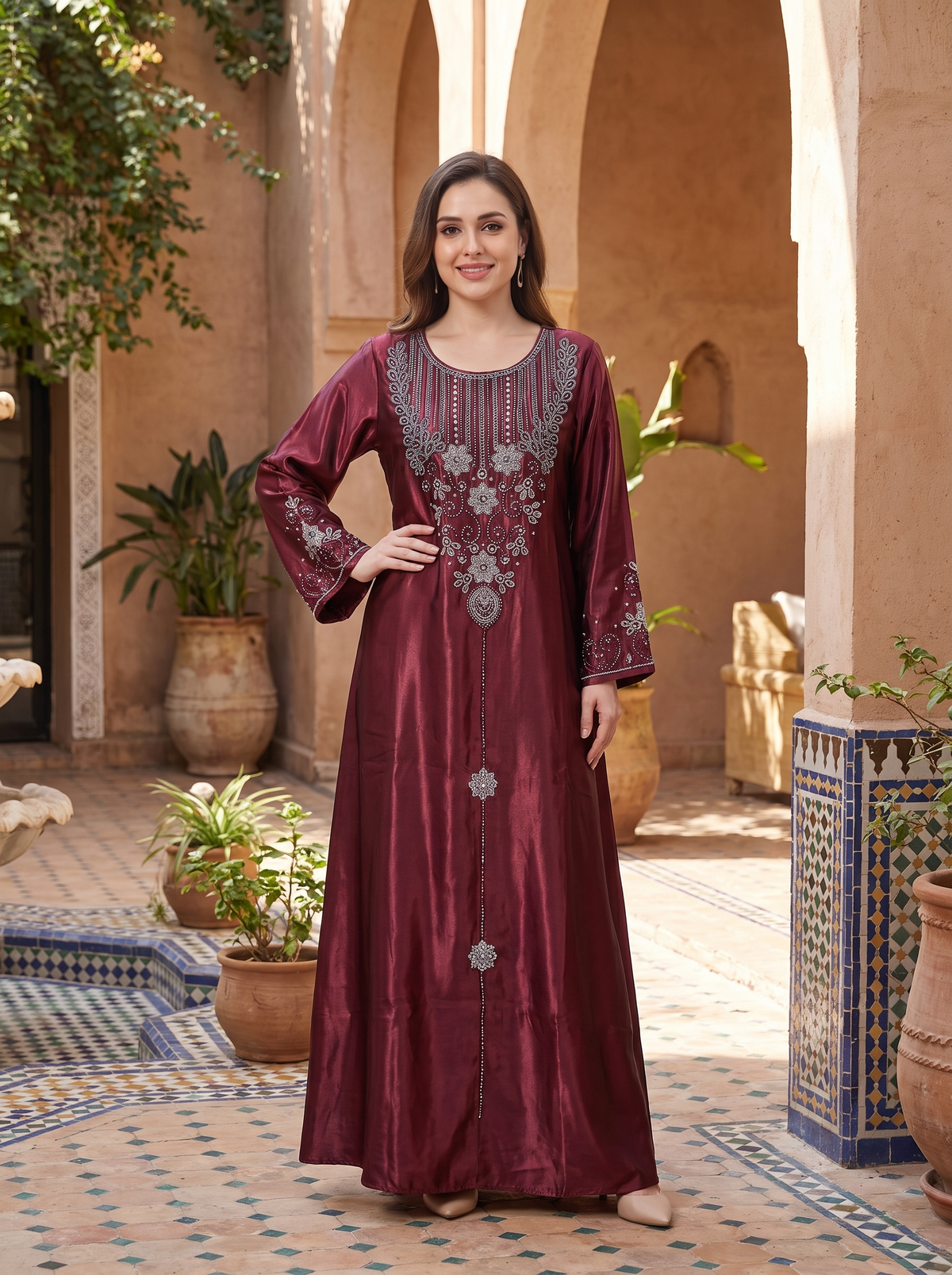 A woman stands in a sunlit courtyard wearing a long maroon satin kurta with silver floral embroidery around the neckline and down the front. The setting features warm beige arches, potted plants, and patterned tile flooring.